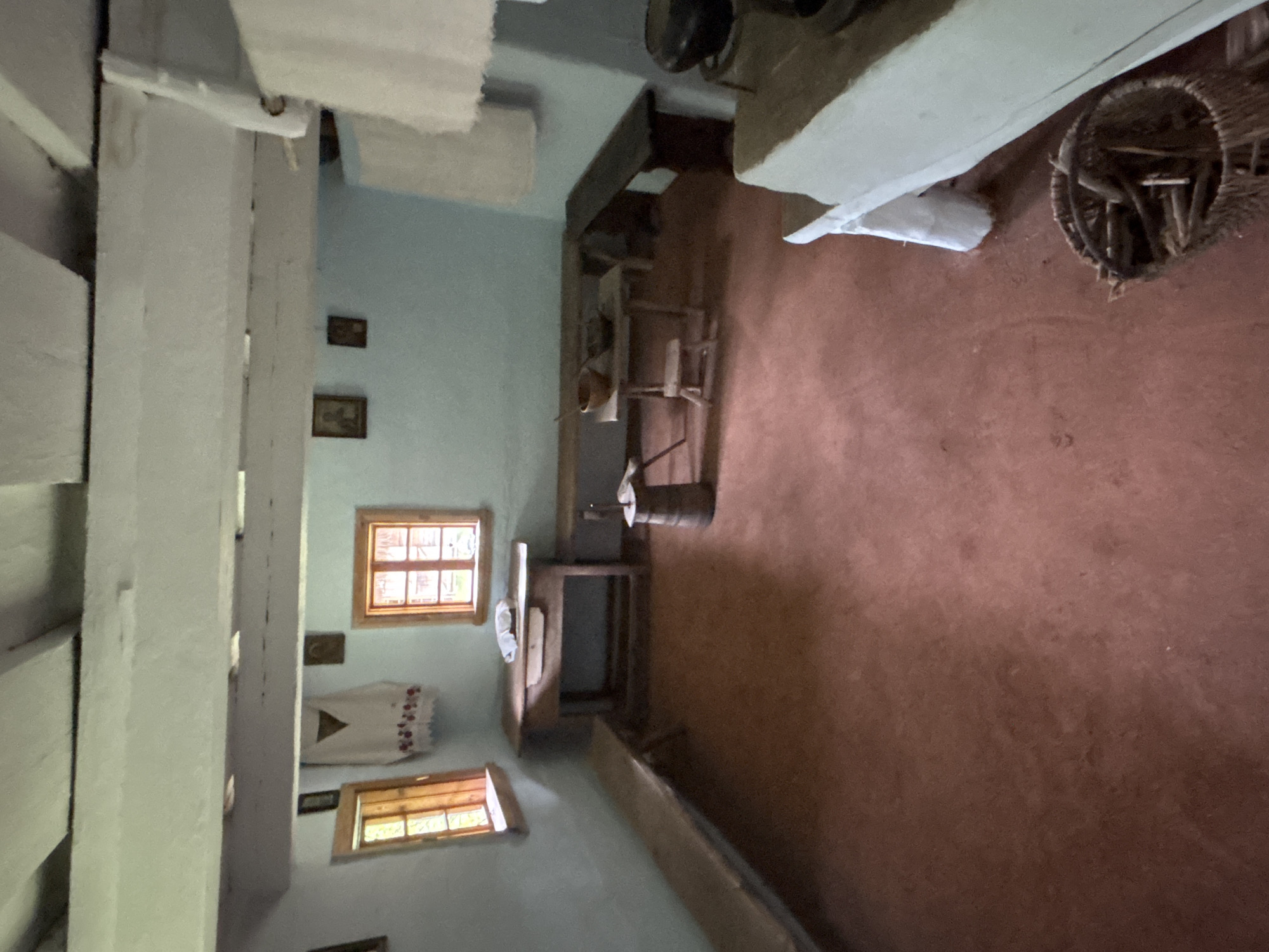 This image depicts the interior of a rustic room, likely a dwelling in Sielachowskie, Poland, given the context. The scene appears to be a historical or traditional setting, possibly a museum exhibit or a preserved old home. The room is sparsely furnished, with an emphasis on functional, rustic objects.
Dominating the space are wooden beams supporting the ceiling and framing the structure of the room. The walls are a light blue, and the floor is a reddish-brown earthen material, suggesting a simple and traditional construction.
Several windows with wooden frames and panes are visible. One larger window on the left shows a grid pattern of smaller panes, letting in natural light. Another smaller window is visible on the right.
Objects in the room include a dark wooden table or work surface with various items on it, including what appears to be a barrel and some metal implements. A basket filled with sticks or kindling sits on the floor near a white object that might be a bag or cloth. There are also framed pictures on the wall, adding a touch of personal history to the space. A piece of embroidered fabric, possibly a wall hanging or tablecloth, with red floral patterns and a triangular motif, is draped over something on the wall.
The lighting suggests it is daytime, with light streaming in through the windows. The overall atmosphere is one of simplicity, history, and perhaps a sense of quietude, evoking a past way of life. There is no visible text in the image.