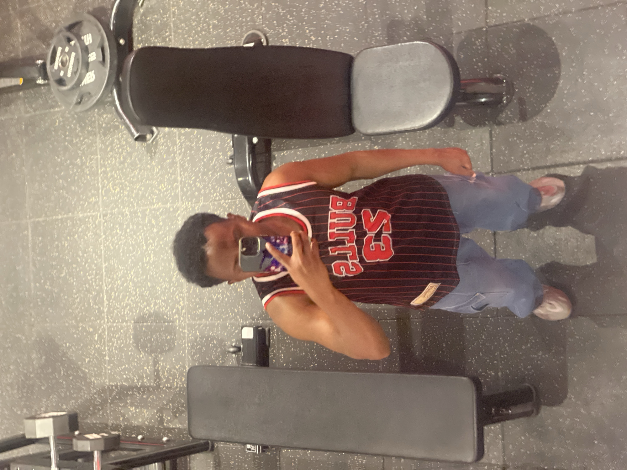 A person is taking a selfie in a gym. They are wearing a Chicago Bulls jersey, light blue jeans, and pink and white shoes. The gym has weight benches and weightlifting equipment. The location is not identifiable as Chongqing High-tech Zone, China.