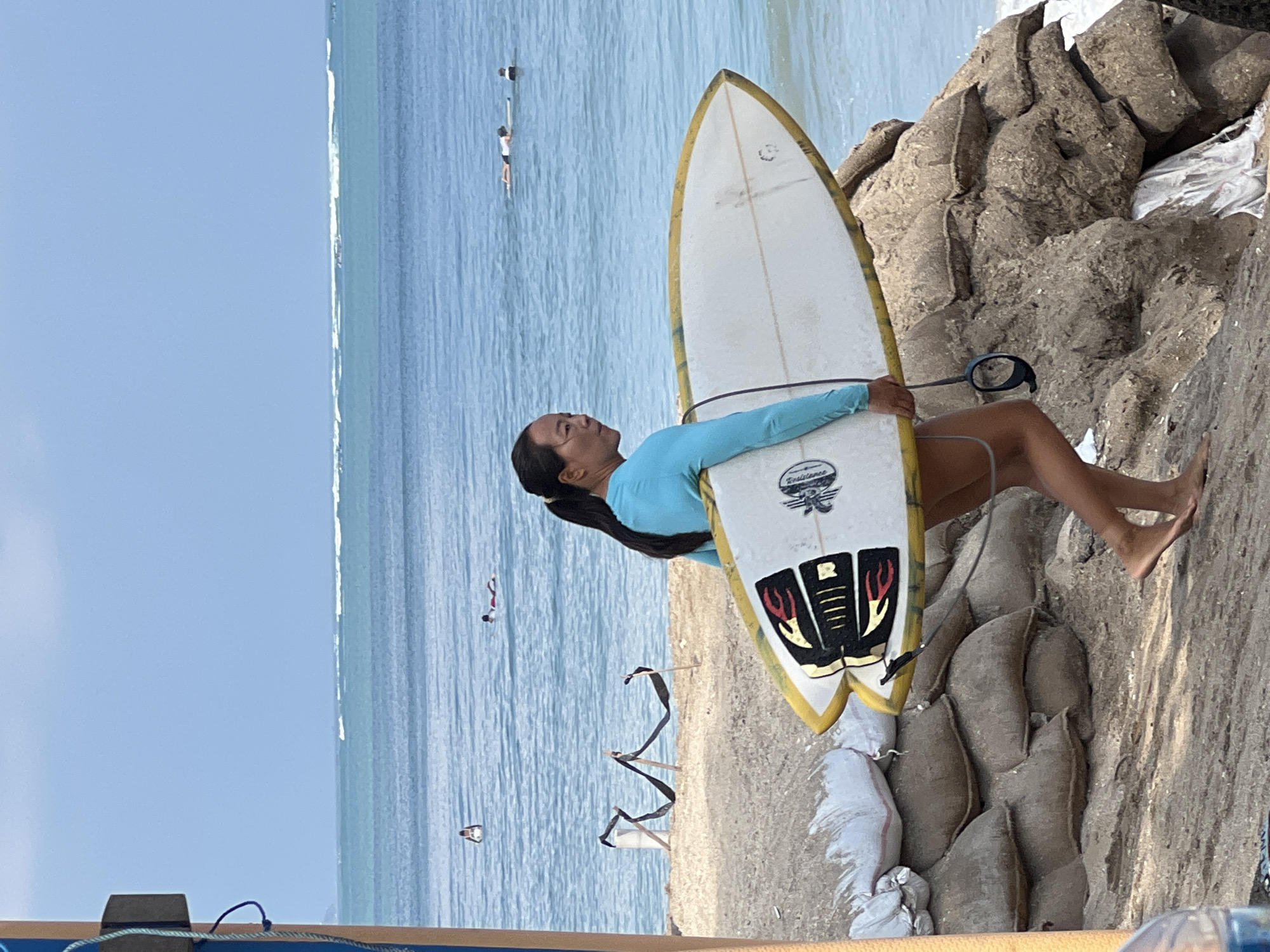 A young Asian woman with dark hair pulled back in a ponytail is walking along a sandy beach, carrying two surfboards under her left arm. She is wearing a long-sleeved teal rash guard and gray shorts. The surfboards are white with yellow rails, and the top one has a graphic design of black fins with red accents. The woman is facing right, looking out at the ocean.
In the background, the ocean is a bright blue, with small white waves breaking in the distance. Several people are visible in the water, some on surfboards and others swimming or floating. The beach itself is sandy, with a line of sandbags piled up along the edge of the water, likely to prevent erosion. The sandbags are a muted brown color and appear to be made of burlap. A dark fabric ribbon is draped between two poles on the beach, near some sandbags. The sky is a clear, pale blue.
The location is identified as Kuta, Indonesia, a popular beach known for its surf breaks and tourist activity. The scene captures a typical day at the beach, with a surfer preparing to enter or leaving the water. The sandbags suggest a protective measure against the sea, hinting at the dynamic nature of the coastal environment in Kuta.