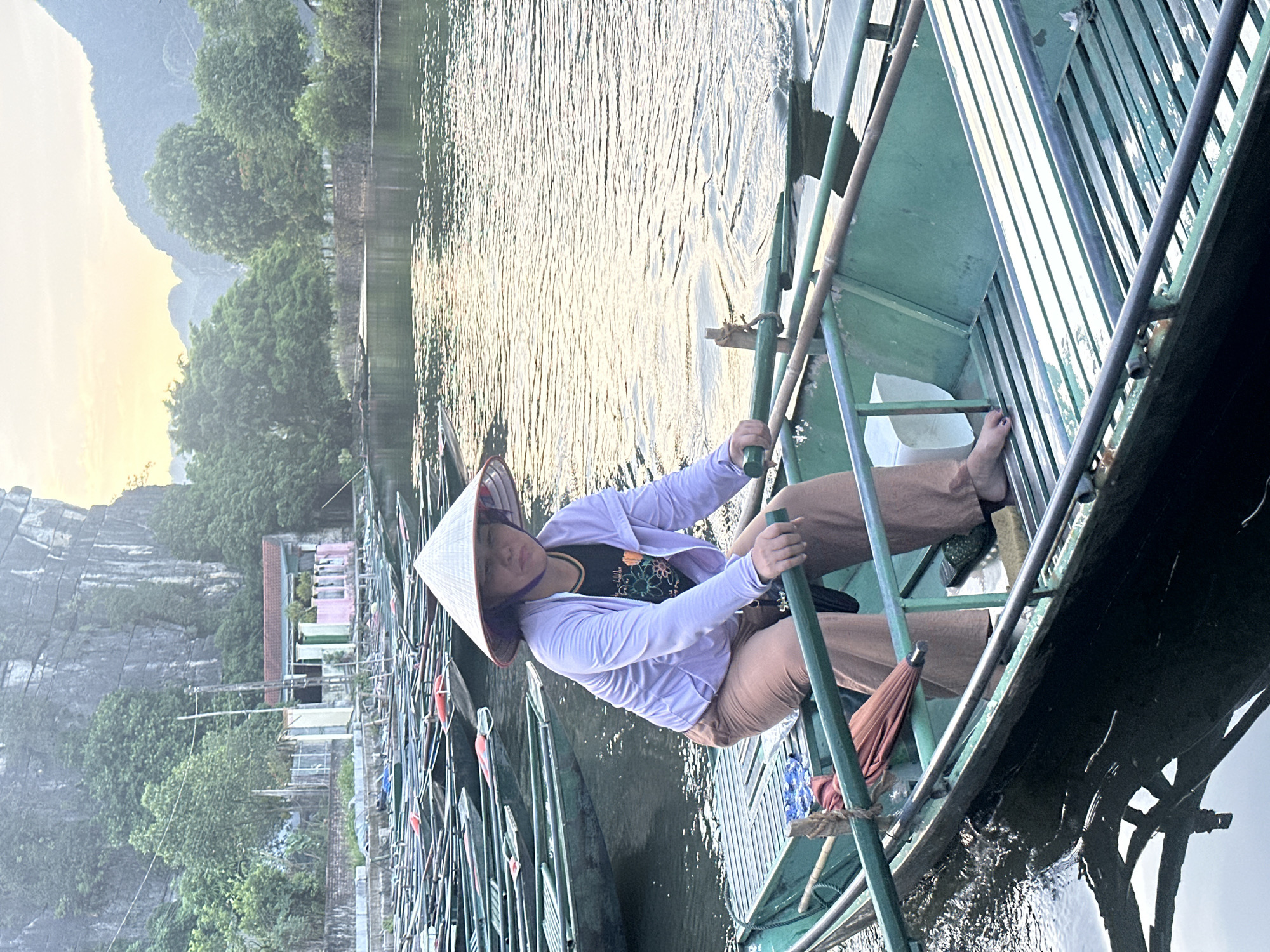 A woman wearing a conical hat and a light purple jacket is seated in a green boat on a river. She is holding oars, suggesting she is rowing. The boat is filled with other similar boats lined up behind it. In the background, lush green trees and rocky formations are visible, characteristic of the landscape in Nam Hoa Lư Ward, Vietnam. The water is calm with ripples, and the sky is overcast with a hint of sunrise or sunset glow.