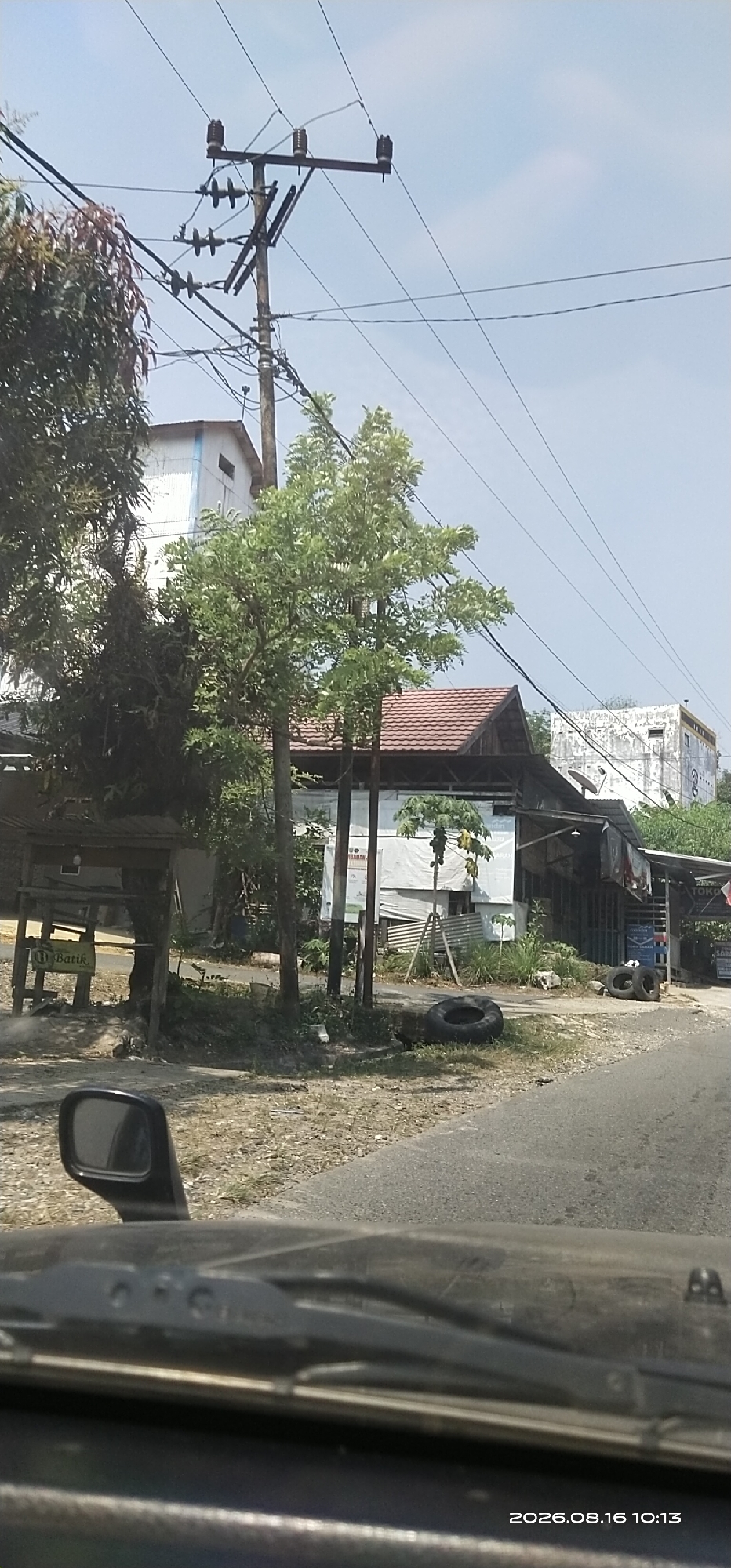 The image captures a roadside scene in Lenggang, Indonesia, under a clear, bright sky. The foreground shows the interior of a vehicle, including a side mirror on the left and the windshield with wipers. The road surface is asphalt, bordered by dry, dusty ground with sparse vegetation.
In the mid-ground, a cluster of buildings is visible. To the left, a small, rustic structure with a corrugated metal roof and a sign that reads "DI BATIK" suggests a shop or stall. Adjacent to it is a tree with dark, dense foliage. Further right, a larger building with a red tile roof appears to be a more substantial establishment, possibly a shop or home, with white sheeting covering part of its exterior. Behind this, taller buildings with white exteriors and some visible windows rise. Electrical infrastructure, including utility poles with multiple insulators and overhead wires, is prominent throughout the scene. Several trees with green leaves are scattered amongst the buildings.
In the background, more buildings are visible, including a white structure with what appears to be a satellite dish. The ground in front of the buildings is rough and unpaved. Several discarded tires are present on the ground.
The timestamp "2026.08.16 10:13" is overlaid in the lower right corner of the image. No people are clearly visible in the scene. The overall impression is one of a rural or semi-urban commercial area in Indonesia on a sunny day.
