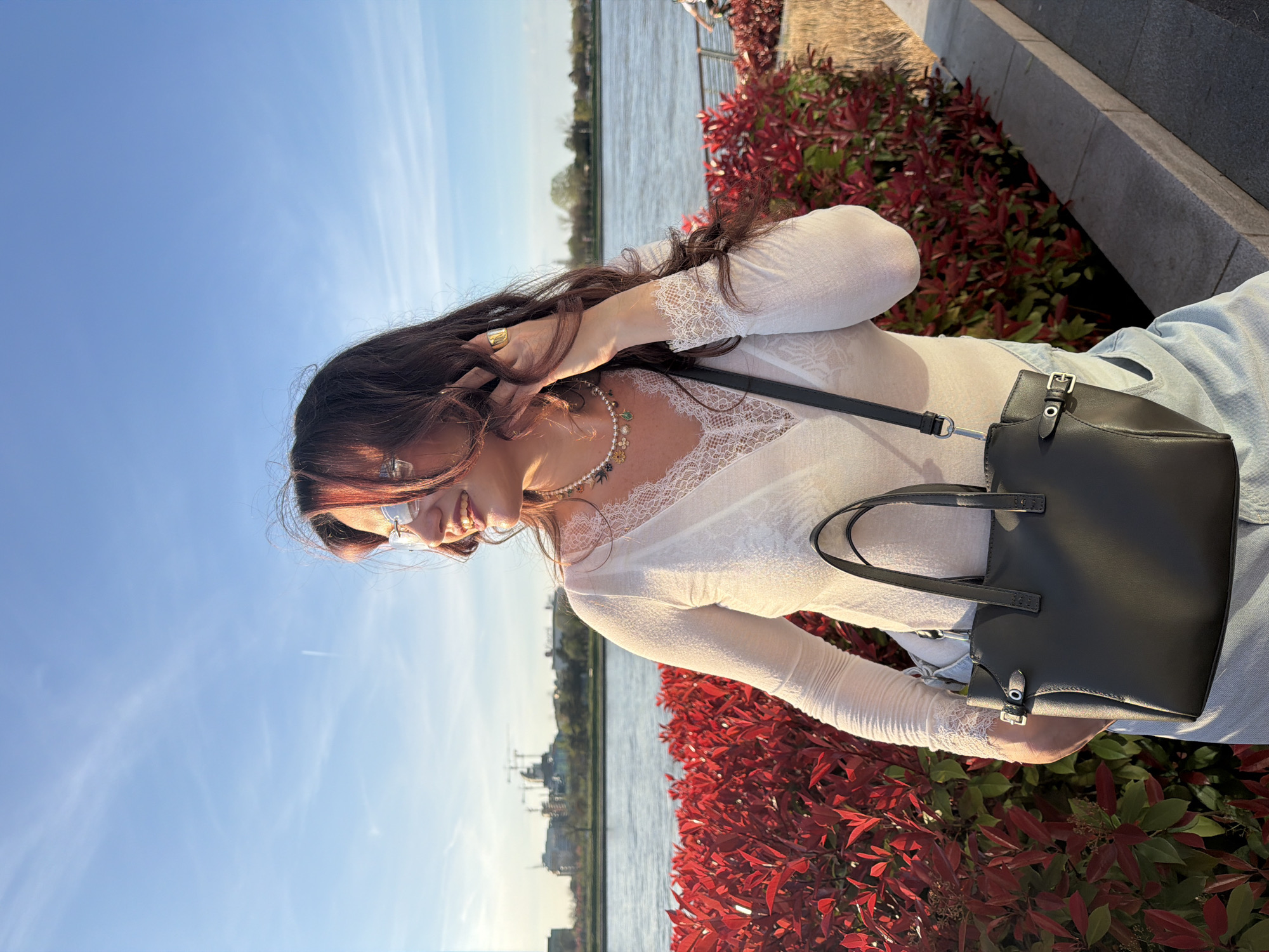 The image depicts a woman outdoors, likely in Belgrade, Serbia, given the urban waterfront landscape in the background. The woman has dark hair with reddish highlights and is wearing silver-rimmed glasses, a white lace-trimmed top, and a pearl necklace. She wears a gold ring on her left hand. She has a black leather handbag with a shoulder strap.
The background features a body of water with buildings visible across the water, indicating an urban setting. To the right of the woman, there are plants with red leaves. The scene is lit by sunlight, suggesting it is daytime.