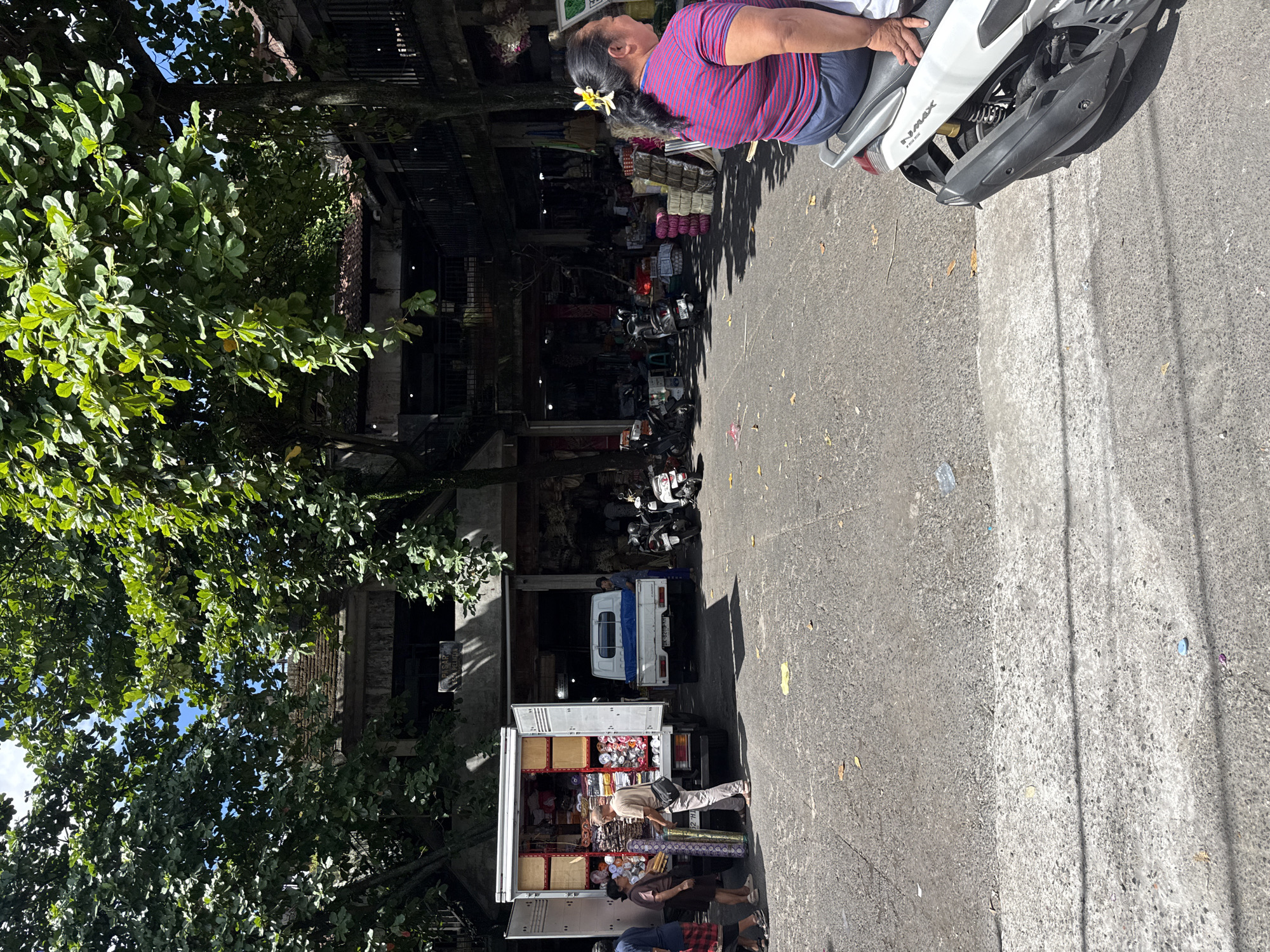The image depicts a street scene in Ubud District, Indonesia. A woman with a flower in her hair sits on a white scooter. A white delivery truck is parked with its back doors open, revealing rolls of textiles inside. People are standing around the truck. Motorbikes are parked in the background near a multi-story building. A tree with green foliage stands on the left side of the image. The road is paved with gray asphalt.
