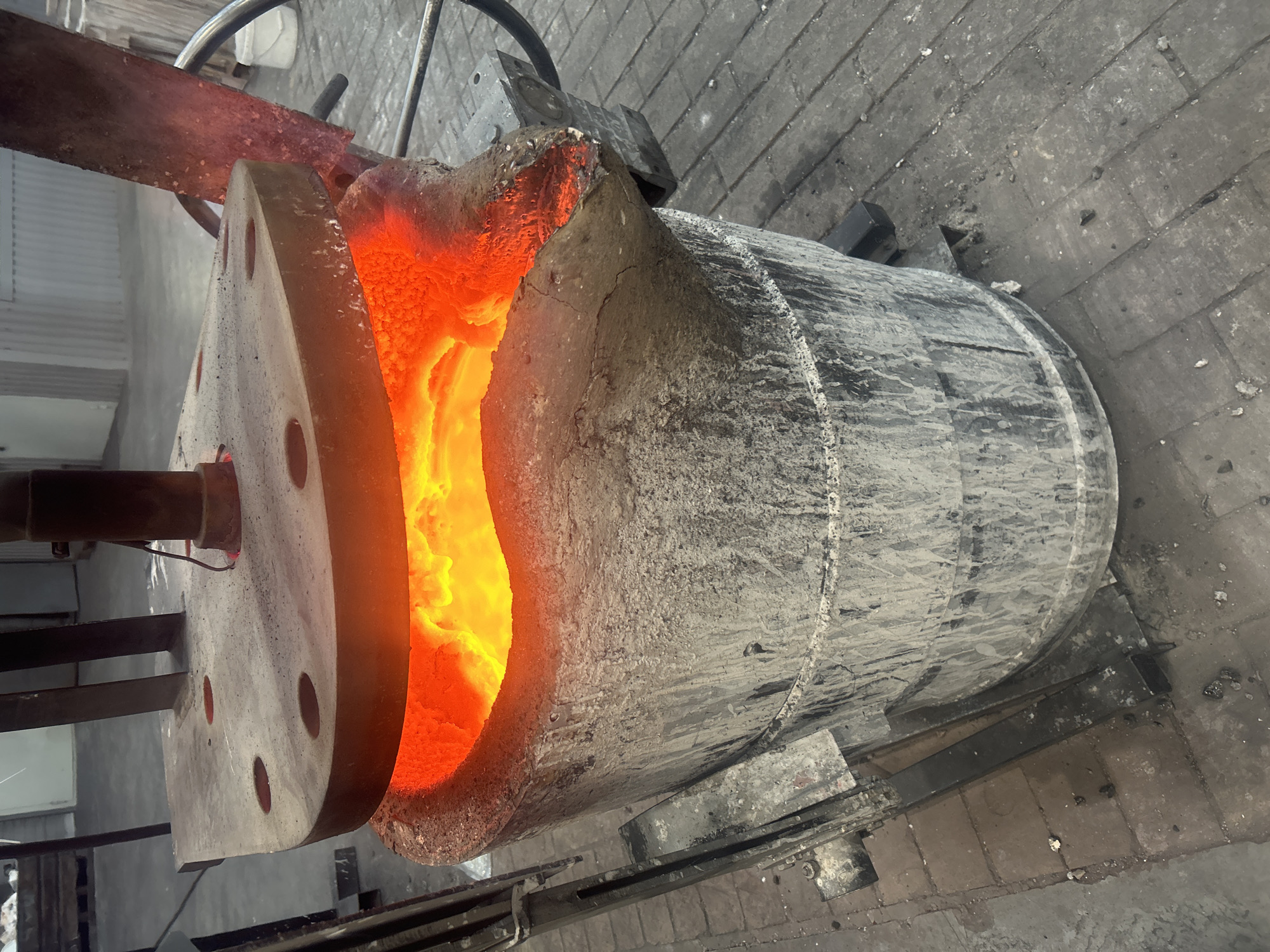 A large crucible, filled with molten metal that glows orange, rests on the floor of what appears to be an industrial setting. The crucible is cylindrical, coated in a rough, grey, possibly heat-resistant material. A metal flange with bolt holes extends from the front of the crucible. A handle or lifting arm is partially visible above the crucible. The floor is tiled with grey bricks. The precise location within the city of Noviy, Russia, is not determinable from the image. No people are visible.