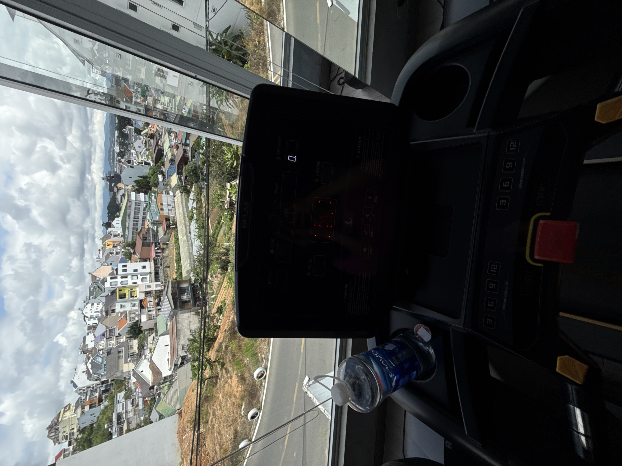 The image shows a treadmill in the foreground with a view of the city of Da Lat, Vietnam, through a large, angled window. The city is built on a hillside, with many colorful houses and buildings clustered together. The sky is partly cloudy. On the treadmill console, a digital display shows "0." A bottle of water is in a holder on the treadmill. The environment appears to be an indoor gym with a view.