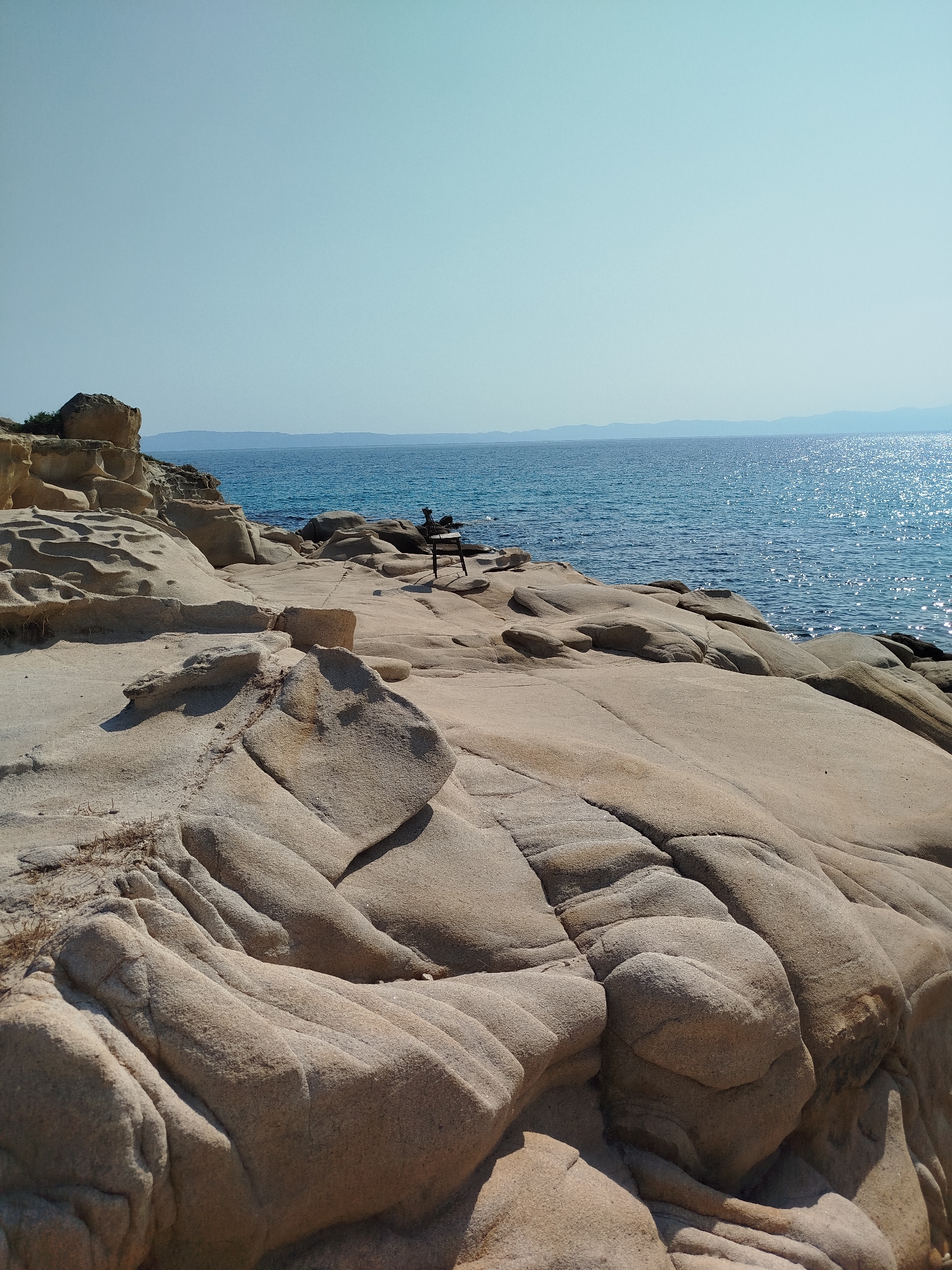 A wide shot captures a sun-drenched rocky coastline with the Aegean Sea stretching out under a clear blue sky. The foreground is dominated by smooth, light-brown rock formations, weathered by time and sea, displaying rippled textures and deep crevices. In the mid-ground, a dark, metal beach chair sits empty on the rocks, facing the water. The sea is a vibrant blue, sparkling with sunlight, and the distant horizon is marked by a faint outline of mountains. The image appears to be taken from a slightly elevated perspective, looking out towards the sea. The location is identified as a rocky outcrop near the settlement of Vourvourou in Sithonia, Halkidiki, Greece, a popular coastal tourist destination known for its natural beauty.