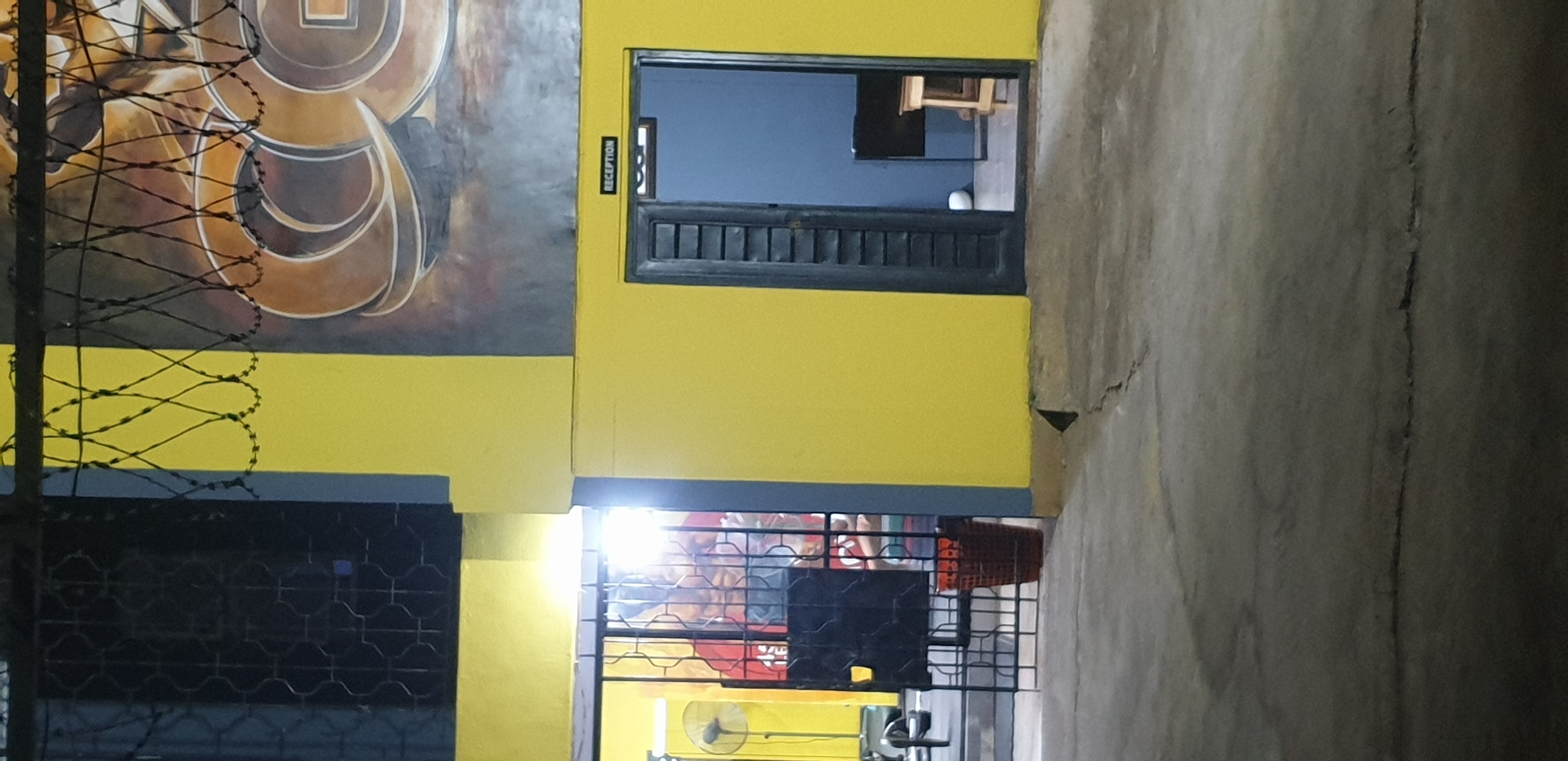 The image captures the exterior of a building in Narayi, Nigeria, painted in vibrant yellow with a grey trim. A large mural, possibly depicting stylized lettering or abstract art, adorns the upper section of the building, partially obscured by a barbed wire fence. To the left, a gated entrance leads into what appears to be a gym or training facility, with a floor fan visible and a person silhouetted behind the bars. A red woven basket sits outside the gate. Further to the right, a door labeled "RECEPTION" is open, revealing a glimpse of a blue-walled interior with a desk and stool. The foreground shows a cracked concrete ground, typical of an urban environment. The lighting suggests it is either dusk or night, with artificial lights illuminating the entrance and reception area.
