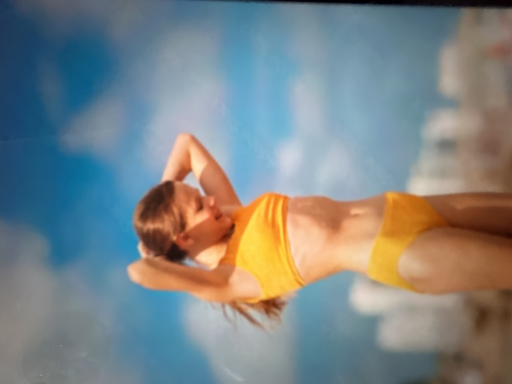 The image depicts a young woman lying down, appearing to be relaxing outdoors. She is wearing a bright orange bikini, consisting of a halter-neck top and matching bottoms. Her head is tilted back, resting on her arms which are clasped behind her head, suggesting a pose of leisure or sunbathing. Her hair is brown and falls around her shoulders.
The background is out of focus, but it appears to be a bright, possibly sunny day with a blue sky and white clouds. There are also some blurred, light-colored shapes in the background that could represent buildings or natural formations, but they are not distinct enough to identify. The overall impression is one of a warm, pleasant environment, perhaps a beach or a poolside, given the woman's attire. There is no visible text in the image.