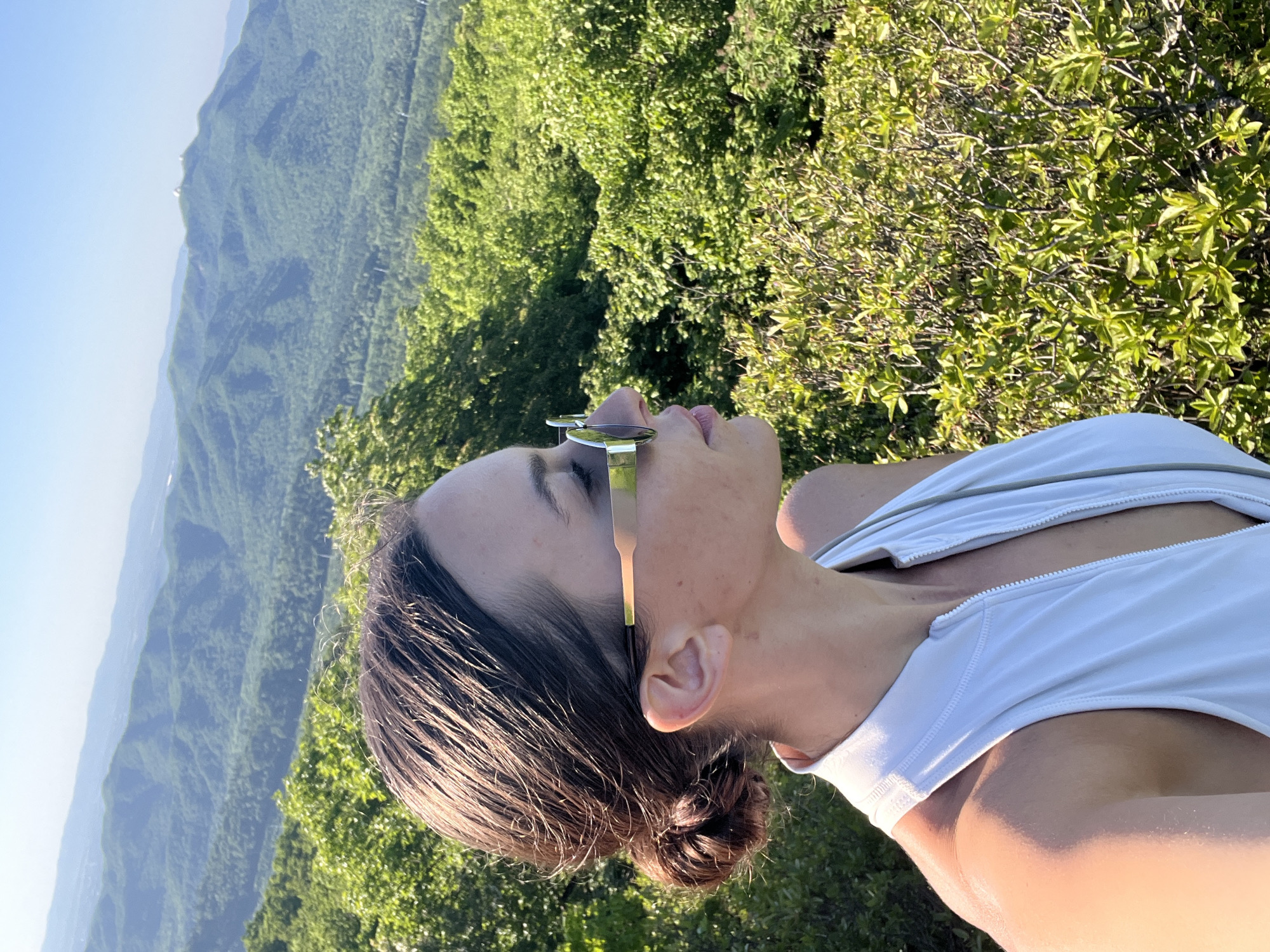 A woman is seen from a low angle, appearing to be looking upward while wearing sunglasses and a white tank top or sports bra. Her hair is pulled back in a bun. The background features a landscape with green vegetation and hills, possibly a mountainous region. The image is rotated slightly, and the lighting suggests it was taken outdoors during the day. It is not possible to determine the location as Vladivostok, Russia from this image.