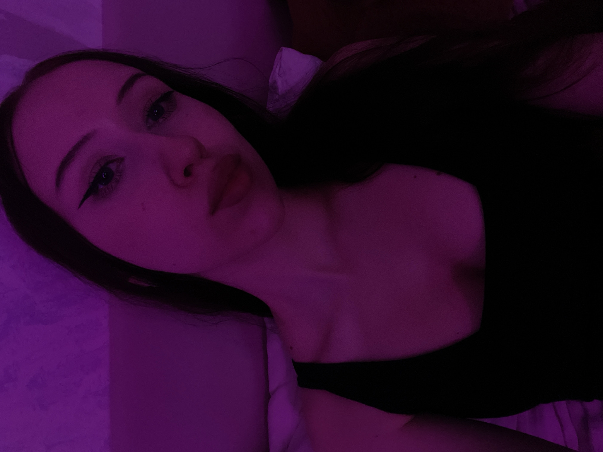 A young woman with long dark hair and winged eyeliner is pictured in a close-up, selfie-style shot. She is wearing a black tank top. The image is lit with a strong purple light, creating a dramatic and atmospheric effect. The background appears to be a textured wall, possibly part of a bedroom or indoor space. There are no identifiable landmarks or contextual clues to place this image in Votkinsk, Russia, or any specific city. The scene is static and focuses solely on the woman's portrait.