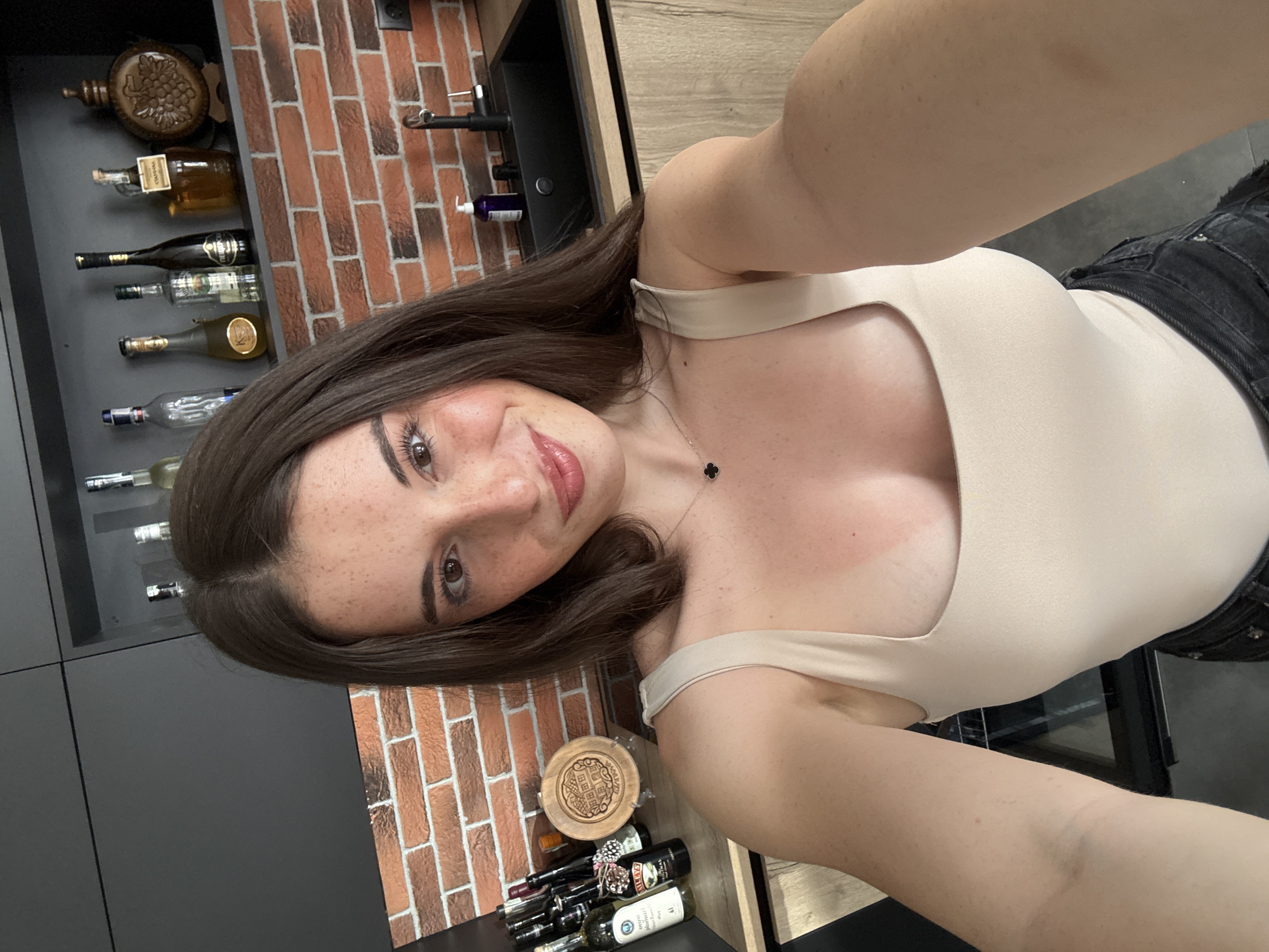 A young woman with brown hair and freckles takes a selfie in a modern kitchen. She is wearing a beige tank top and black shorts. The kitchen features dark grey cabinets above a brick backsplash, and light wood cabinets below a black sink and faucet. On the shelves above the sink are several bottles, and on the counter to the left are more bottles and a wooden plate. The woman's arm is extended, holding the camera.