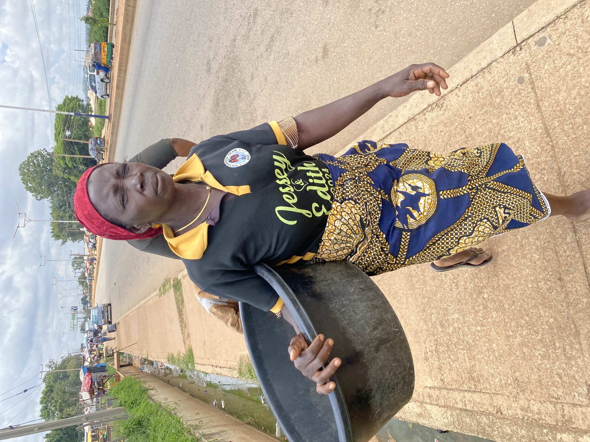 In Bukuru, Nigeria, a woman wearing a red headscarf and a black polo shirt with yellow trim is walking on a sidewalk, carrying a large black basin. Her shirt has the text "Jessey & Edith 2024" printed on it, along with a circular logo. She is also wearing a patterned blue and yellow wrap skirt and sandals. The sidewalk is made of concrete and is adjacent to a busy road with traffic, including cars and a motorcycle taxi. On the other side of the sidewalk is a drainage ditch filled with murky water and debris. In the background, there are market stalls with red and white awnings, trees, and more vehicles on the road. The sky is overcast with clouds.