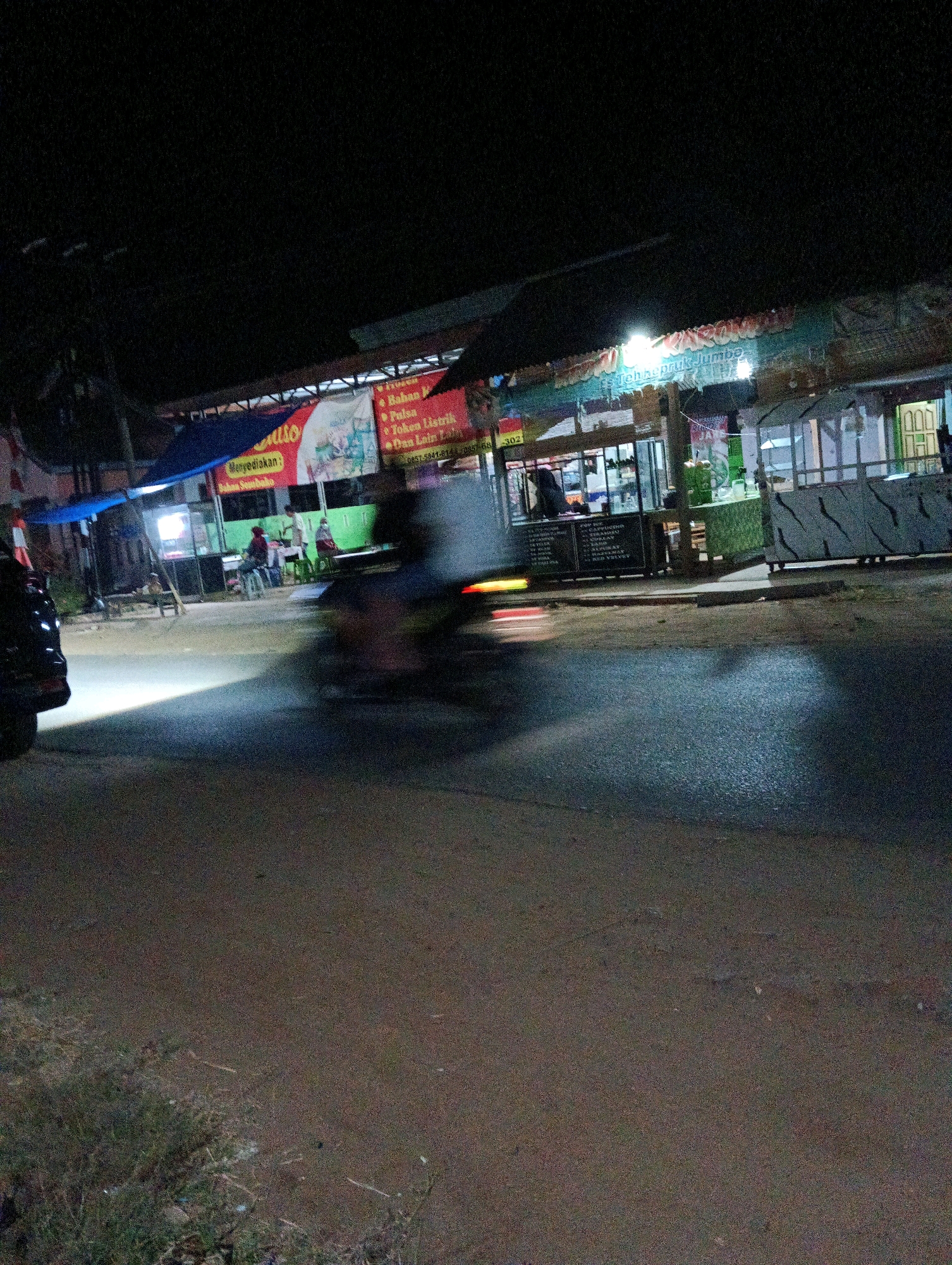 The image depicts a nighttime street scene in Adirejo, Indonesia. A motorcycle is in motion in the foreground, creating a blur of motion. In the background, several food stalls are visible, illuminated by bright lights. One stall has a sign that reads "Teh Kepriik Jumbo," indicating it sells jumbo tea. Other signs mention "Bahan P," "Pulsa," "Joken Listrik," and "Dan Lain Lain," suggesting a variety of goods and services are offered. There are people visible at the stalls, some standing and some seated. A black vehicle is parked on the left side of the image. The environment appears to be a roadside with a paved street and some vegetation on the side.