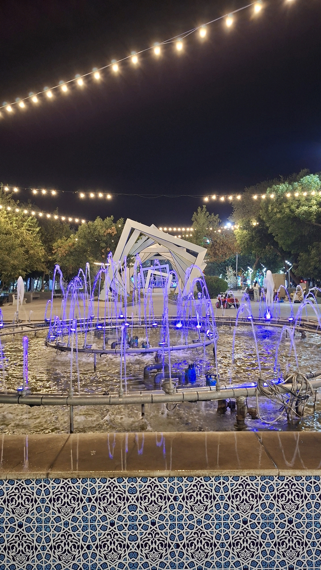 An outdoor fountain with purple lights is illuminated at night in a city park in Navoiy City, Uzbekistan. The fountain is in the foreground, with water jets spraying upwards. In the background, there is a modern white art installation with geometric shapes and trees with string lights overhead. The scene is illuminated by the artificial lights of the fountain, string lights, and possibly streetlights. No people are clearly visible in the immediate vicinity of the fountain. The lower part of the image shows a tiled border with a geometric pattern in blue and white.