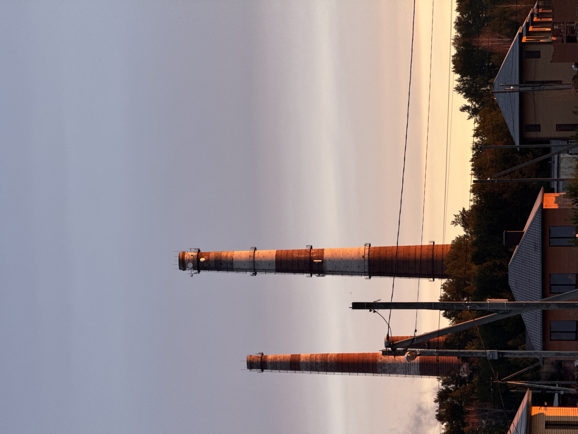 The image shows two industrial chimneys in Chaikovsky, Russia. The chimneys are red and white striped, with metal supports and platforms near the top of the taller chimney. They are positioned against a backdrop of a pale, clear sky transitioning to a sunset hue. In the background, trees and buildings are visible. Power lines stretch across the scene. The chimneys are a dominant feature, suggesting an industrial area within the city.