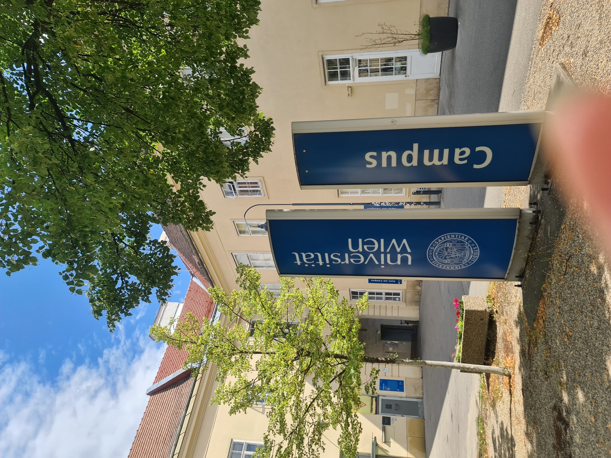 The image shows a sign for "Universität Wien" and "Campus" in front of a cream-colored building with red tile roofs. The building has several windows, some of which are open. A tree with green leaves is planted in front of the building. A planter with pink flowers is visible in the foreground. The sky is blue with some white clouds. The scene appears to be an outdoor area on a university campus.