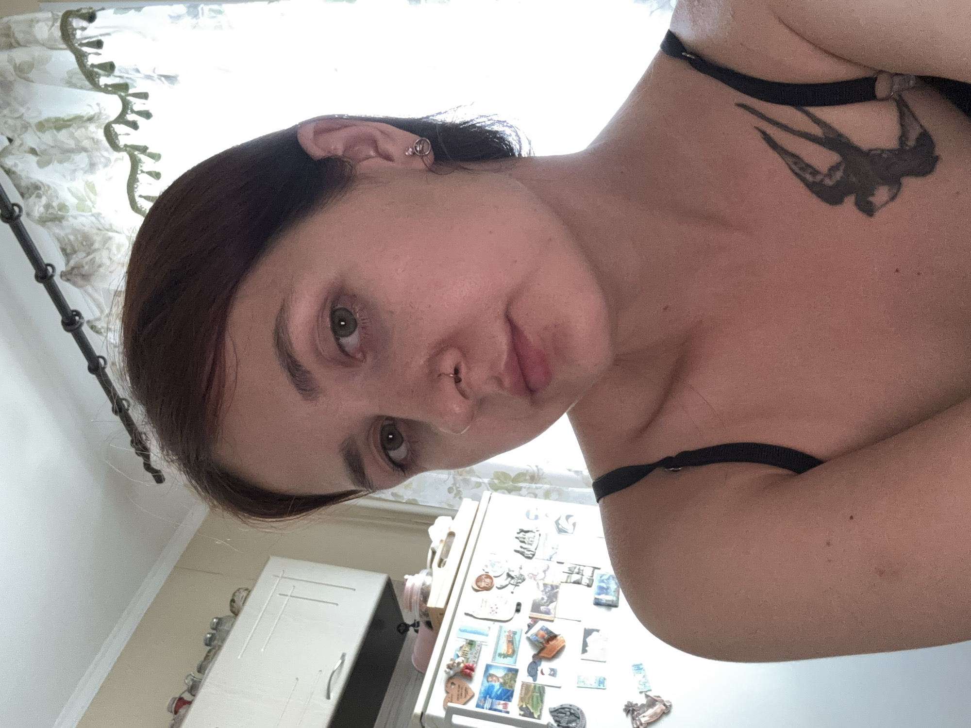 A medium shot portrait of a young woman with short, dark brown hair and light green eyes. She is looking directly at the camera with a neutral expression. She has a nose ring and a small silver earring in her right ear. She is wearing a black spaghetti strap top, and a tattoo of a swallow is visible on her left shoulder. The background appears to be a kitchen, with white cabinets and a refrigerator visible on the left. The refrigerator is decorated with numerous magnets and photographs. A window with a floral curtain is visible on the right. The lighting suggests it is daytime, with natural light coming from the window. The location is described as a kitchen in Naberezhnye Chelny, Russia.