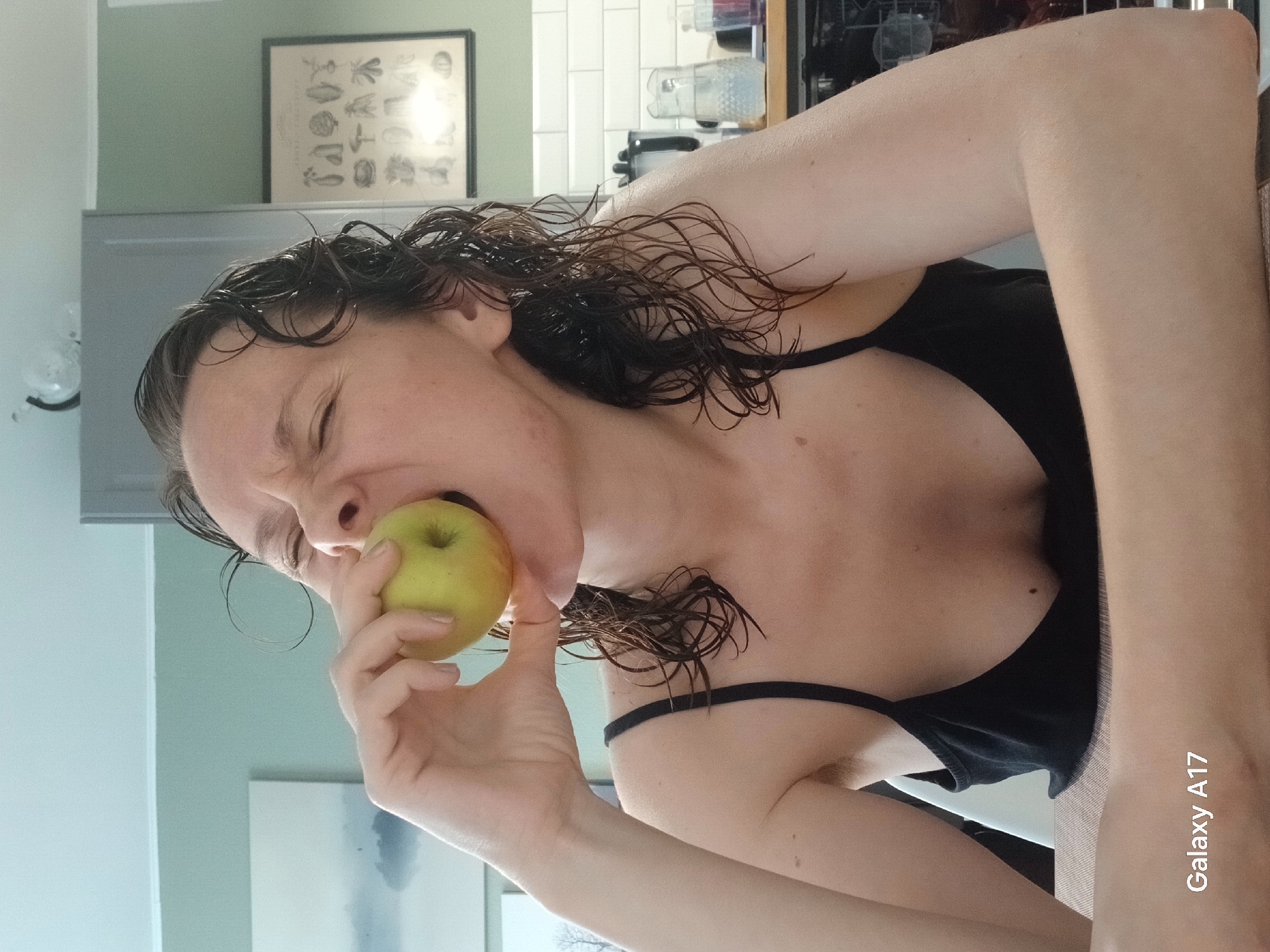 The image depicts a woman indoors, biting into a green apple. She is fair-skinned with wet, dark, curly hair. She is wearing a black tank top. The background shows a kitchen setting with white tile backsplash and a framed piece of artwork above a light-colored countertop. A "Galaxy A17" watermark is visible in the lower right corner of the image. The location in the city of Yekaterinburg, Russia, cannot be determined from the image.