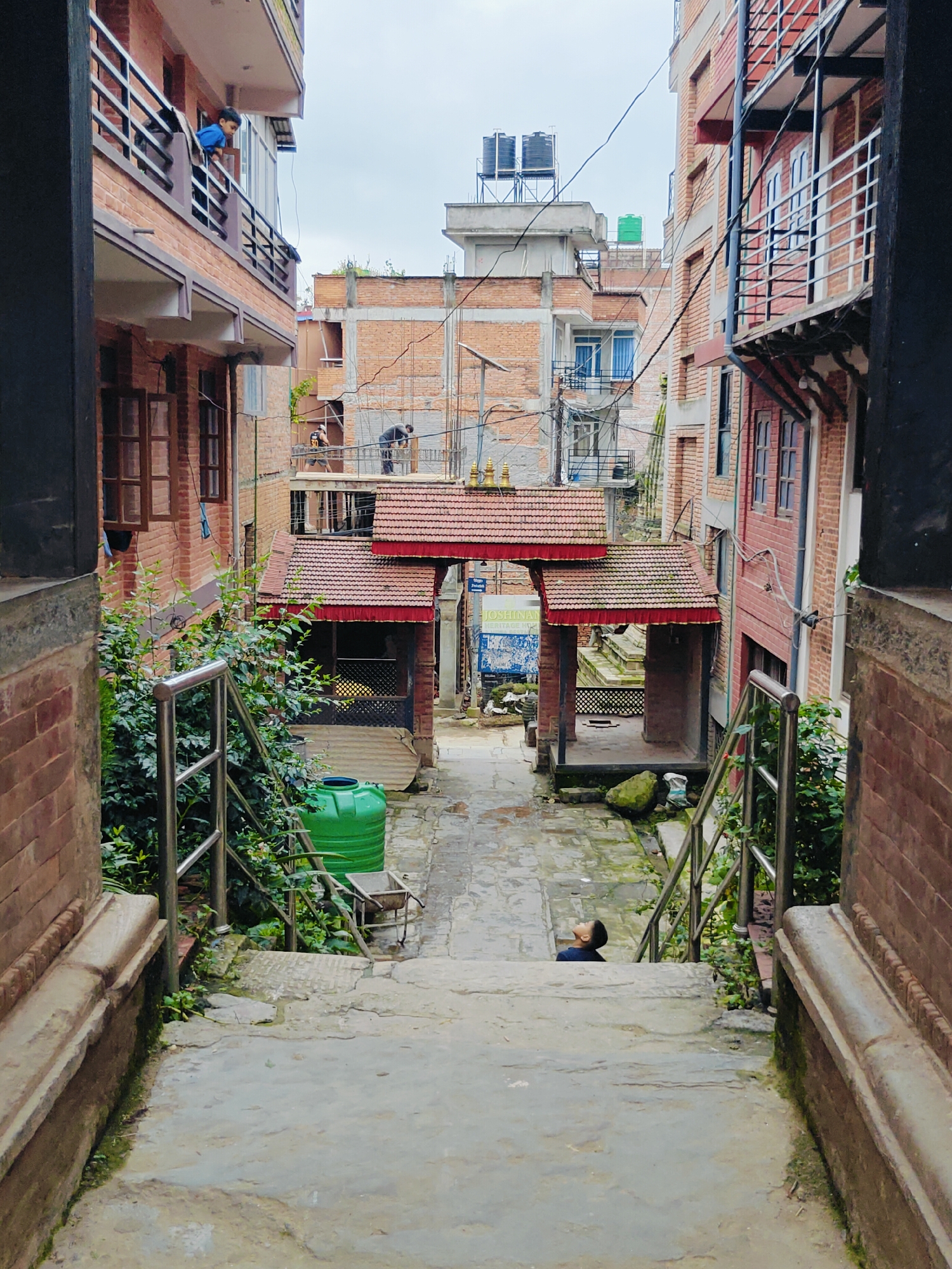 The image depicts a narrow alleyway in Kirtipur, Nepal, lined with brick buildings. In the foreground, a set of concrete stairs descends into the alley. On the left side of the stairs, a thick patch of greenery grows, with a metal railing bordering the stairs. A large green water tank is situated to the right of the greenery.
Further down the alley, a traditional gateway with a red-tiled roof, supported by wooden columns, spans the path. Beyond the gateway, the alley continues, with more brick buildings visible on either side. A young boy wearing a dark blue shirt is lying on the stairs looking upwards, his head propped on his hands.
In the upper floors of the buildings, balconies are visible, with some people standing or leaning over the railings. The sky is overcast. The overall scene suggests a residential or mixed-use area within a Nepalese city.