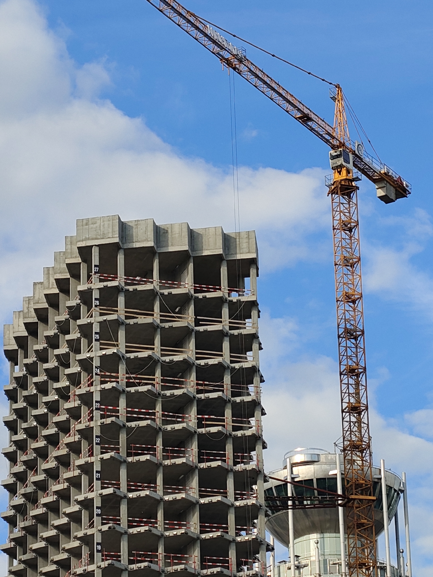 The image depicts a construction site in Moscow, Russia, featuring an unfinished concrete building alongside a tall, orange construction crane. The concrete building, identifiable by its floor numbers labeled from 19 to 27, has a modular design with open spaces. A distinct structure with a metallic, disc-shaped element rests near the base of the construction crane and the concrete building. The backdrop showcases a blue sky with scattered white clouds.