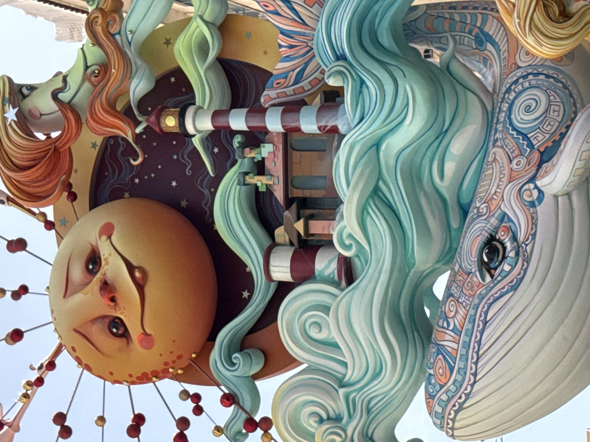 This is a close-up, low-angle shot of an elaborate and colorful artistic installation, likely a sculpture or float, against a bright, clear sky. The installation features several whimsical, stylized figures and objects.
Dominating the lower left of the frame is a large, round, yellow face resembling the sun, with a human-like expression. It has large, dark, somewhat sad-looking eyes, rosy cheeks, and a painted mouth with a downturned expression. Red and gold beads hang from strings attached to this face, creating a festive, almost mobile-like effect.
To the right of the sun face, and extending upwards, is a large, pale blue-grey creature that appears to be a whale. Its head is visible, with intricate, swirling patterns in shades of blue, orange, and white decorating its face around a dark eye. The whale's form is smooth and flowing.
Interspersed between these main figures are abstract, organic shapes, primarily in light green and a vibrant turquoise. These shapes, with their swirling and flowing lines, seem to mimic waves or natural forms, adding to the dynamic and dreamlike quality of the installation.
A red and white striped object, resembling a telescope or a piece of a lighthouse, is also visible, adding a touch of linear design amidst the more organic curves. The background consists of a dark circular area with small white stars, possibly representing the night sky, and beyond that, a bright, clear sky, suggesting the installation is outdoors during the daytime.
The overall impression is one of fantasy and celebration, with a vibrant color palette and playful, almost surreal imagery. There is no visible text in the image.
