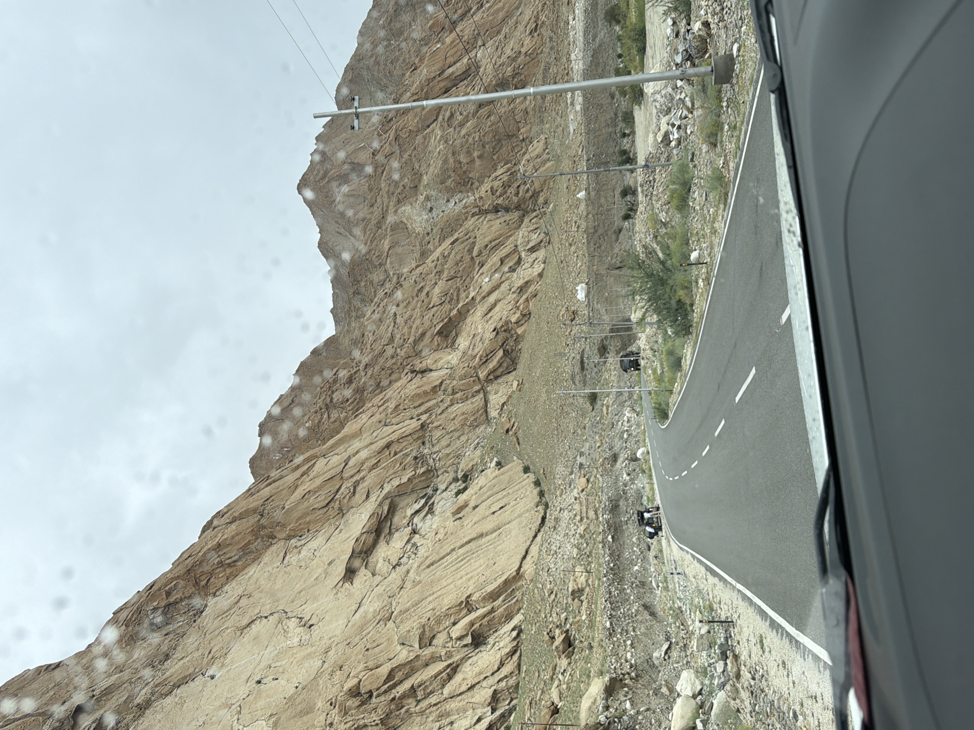 The image shows a winding road in a mountainous region. The road curves to the right and is flanked by dry, rocky terrain and steep, tan-colored mountains on the left. The sky is overcast and a light gray. Water droplets are visible on the windshield, suggesting it might be raining or has recently rained.
Several objects are present along the roadside:
* **Vehicles:** Two small vehicles are visible on the road. One, a dark-colored SUV or jeep, is parked on the right side of the road further ahead. Another vehicle, appearing to be a motorcycle with a sidecar or a small utility vehicle, is stopped on the left side of the road. Two people are standing near this vehicle, seemingly attending to it.
* **Infrastructure:** Several utility poles with wires are scattered along the sides of the road. Small orange and white markers are also visible along the edge of the road.
* **Vegetation:** Sparse, dry vegetation, including bushes and grasses, grows in patches along the lower slopes of the mountains and the roadside.
The foreground of the image shows the interior of a vehicle, specifically the dashboard and windshield wipers. The overall scene depicts a journey through a rugged, arid landscape under a cloudy sky.
The location is identified as Likche, India, suggesting a remote, high-altitude area.
