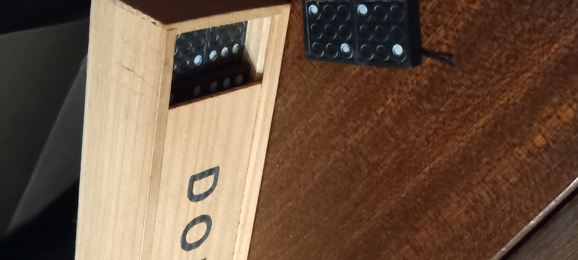 This media file depicts an indoor scene in Kryvyi Rih, Ukraine, featuring a collection of dominoes and their wooden storage box. No people are visible.
The primary subjects are a light-colored wooden box and several black dominoes. The box, positioned vertically on the left side of the frame, has an open top revealing a stack of dominoes inside. The word "DOMINO" is partially visible in dark lettering on the side of the box, with the letters "DO" clearly legible.
To the right, on a textured, reddish-brown surface that appears to be a table or countertop, another set of dominoes is arranged. These are also black with white pips, and they are laid out in a grid-like pattern, possibly part of a game or demonstration. A thin, dark string or cord is visible near this set of dominoes.
The lighting is soft and ambient, suggesting an indoor setting without direct sunlight. The overall impression is one of leisure or a casual setup for a game.