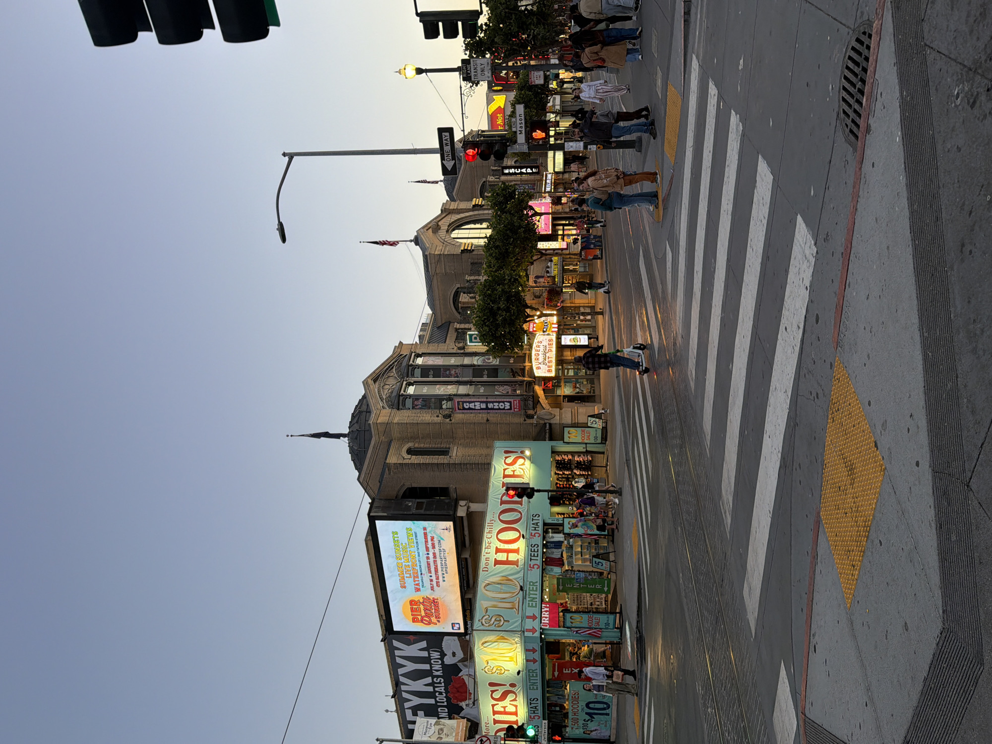 This image captures a street scene in San Francisco, California, likely near Fisherman's Wharf, given the prominent "PIER BIRTHDAY PARTY" advertisement. The time of day appears to be late afternoon or early evening, with soft, warm light illuminating the buildings and street.
In the foreground, a pedestrian crosswalk spans the width of the image, with white stripes marking the pedestrian path. The sidewalk to the right features a tactile paving square, indicating accessibility for visually impaired individuals. Several people are present, some waiting to cross, others walking across the street, and a few are seen on electric scooters.
The buildings lining the street display a vibrant mix of signage. A large digital billboard advertises a "PIER BIRTHDAY PARTY" with "SUMMER SUNSETS, LIVE MUSIC, WATERFRONT VIEWS." Below this, a storefront with brightly lit signs offers "$10 HOODIES!" and "$5 HATS." Other businesses visible include "BURGERS BEST PIES" and a "GAME SHOW" establishment. The architecture of the buildings is varied, with some featuring ornate facades and domes, hinting at historical significance.
Streetlights and traffic signals are visible, with one traffic signal showing a red light. The sky above is a clear, pale blue, transitioning to a lighter hue near the horizon. The overall impression is a bustling urban environment with a mix of commercial activity and pedestrian traffic.