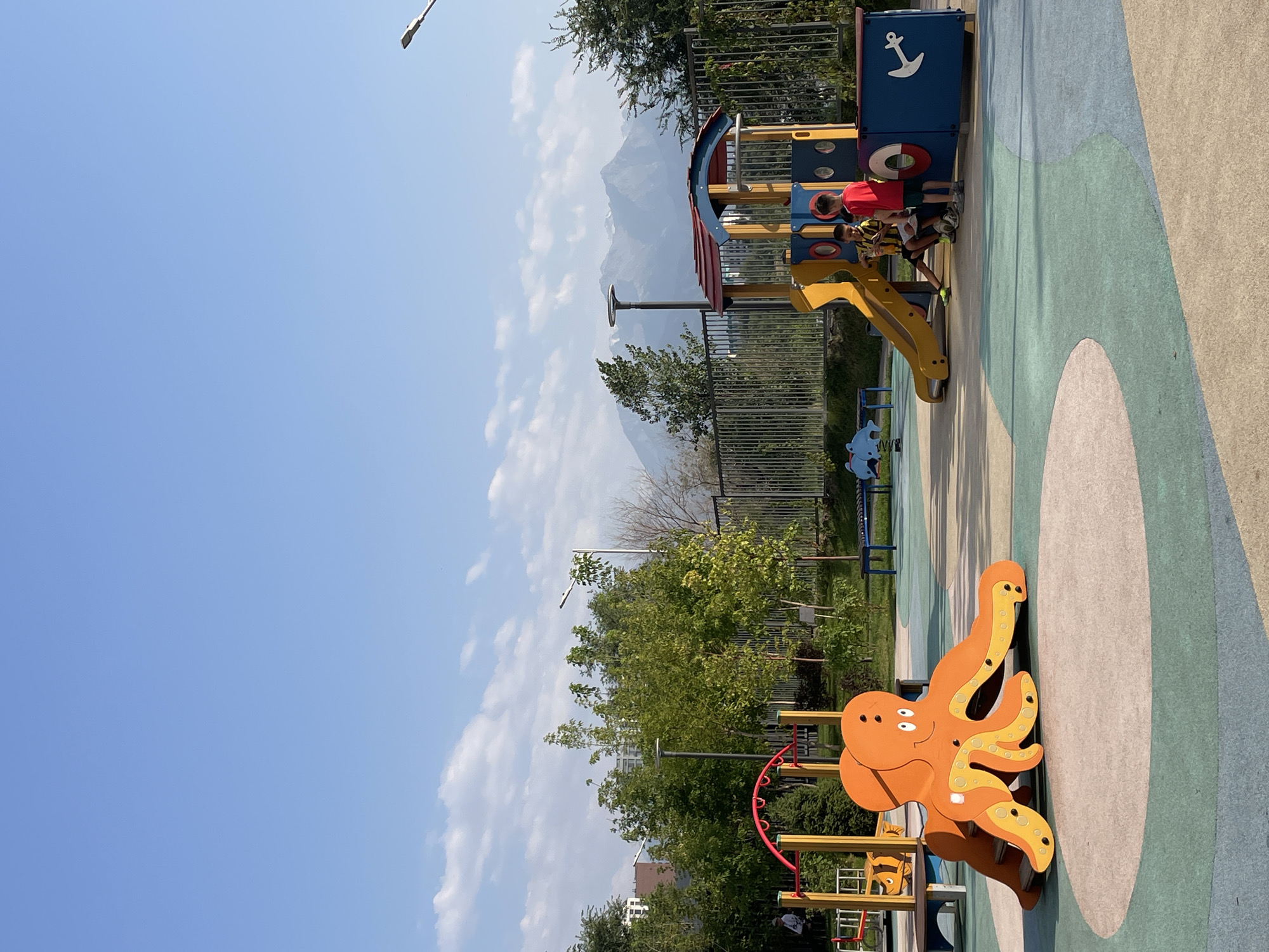 A bright, sunny day is captured on a modern outdoor playground. The ground is a colorful, soft-surface material with large, circular green and beige sections. In the foreground, an orange octopus-shaped climbing structure with a slide emerges from the ground. Behind it, a yellow and red playground set with a slide and a boat-themed section is visible, with two young boys, one in a red shirt and the other in a black and yellow striped shirt, interacting near it. Trees and lush green foliage surround the playground, with a metal fence in the middle ground. In the background, a large, snow-capped mountain range dominates the horizon under a clear blue sky with scattered white clouds. Buildings and streetlights are also visible in the distance. The scene appears to be located in Almaty, Kazakhstan, given the distinctive mountain landscape.