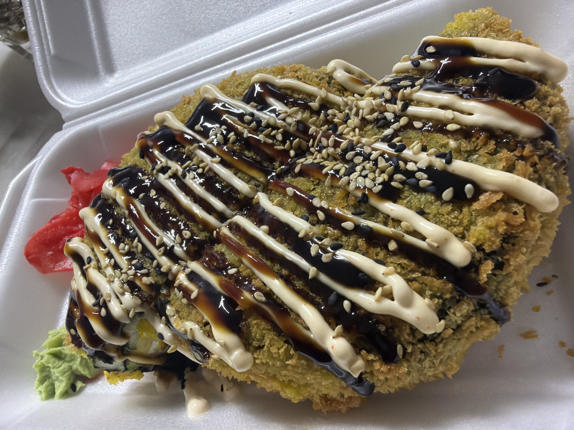 The image displays a close-up, overhead view of a deep-fried sushi roll served in a white styrofoam takeout container. The roll is covered in a green, crispy coating and drizzled with both a dark brown sauce and a creamy white sauce, possibly mayonnaise. Sesame seeds, both black and white, are sprinkled on top. To the upper left of the roll, there is a dollop of bright green wasabi and a few slivers of pink pickled ginger. The background consists of the textured interior of the takeout container. There are no people or actions depicted. The image does not provide any information regarding its location in Strășeni, Moldova.