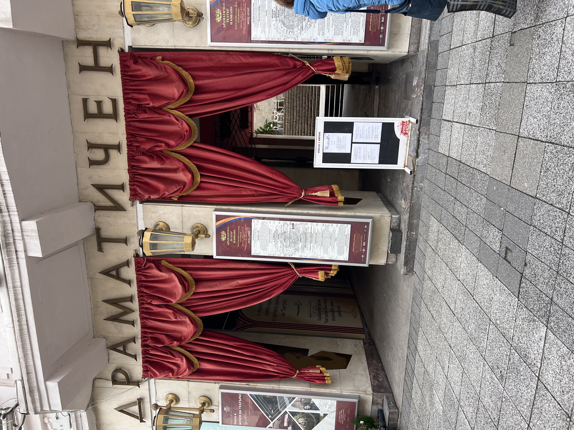 The image shows the entrance to the Plovdiv Drama Theater in Plovdiv, Bulgaria. The entrance is decorated with red velvet curtains and gold tassels. The name of the theater, "ДРАМАТИЧЕН ТЕАТЪР" (Dramatic Theater), is written vertically on the building's facade in bronze letters. There are posters and signs displayed on either side of the entrance, providing information about upcoming performances, online ticket sales, and the theater's museum. A Stella Artois beer advertisement is also present near the entrance. The sidewalk is paved with gray cobblestones. Part of a person dressed in a blue coat is visible in the upper right corner.