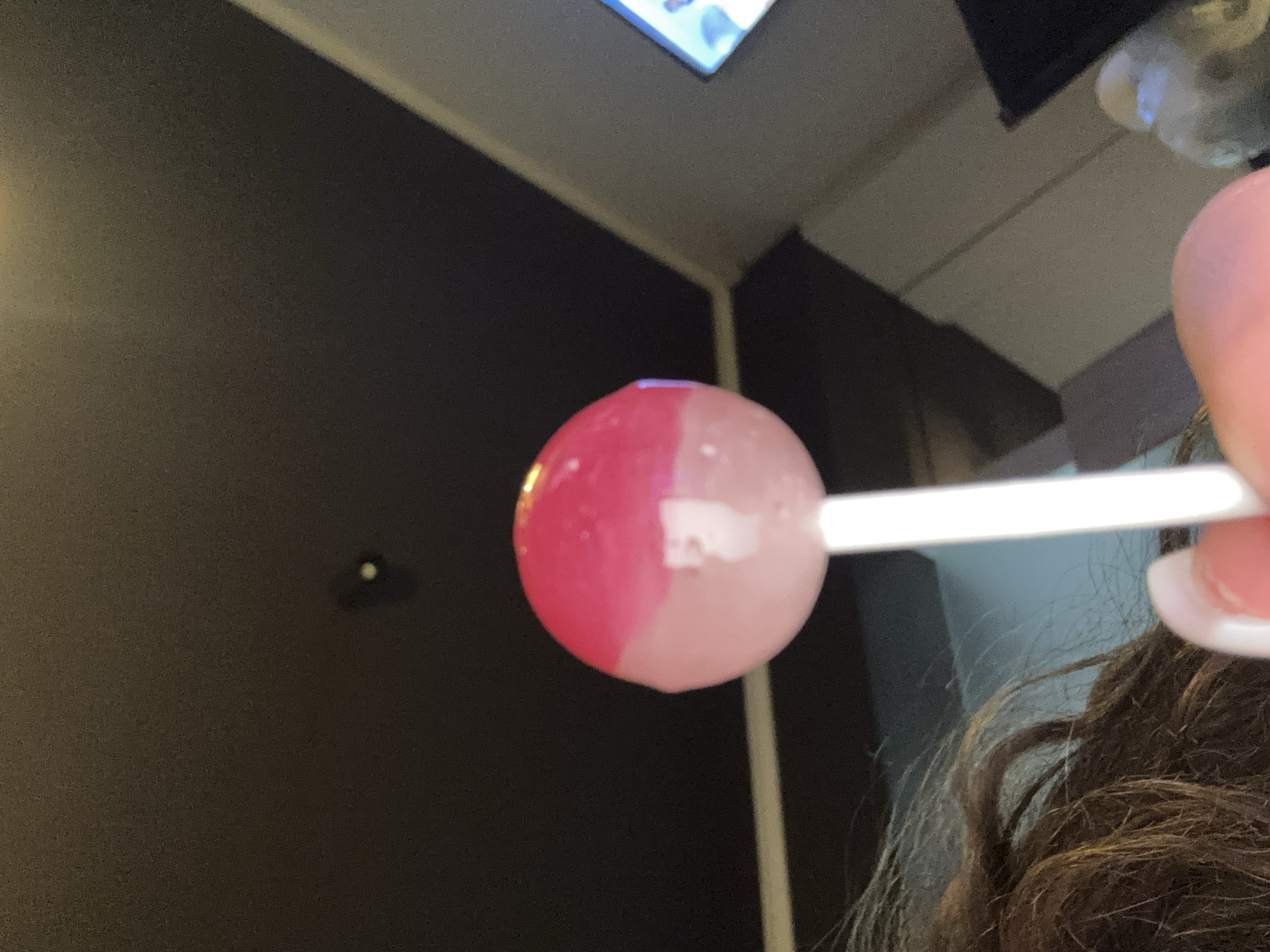 A person is holding a lollipop. The lollipop is round and has two colors, pink and white. The stick is white. The person has long brown hair and white painted nails. The background is dark, with a glimpse of a light-colored ceiling. It is not possible to determine the location of the image within Sofia, Bulgaria, based on the provided information.