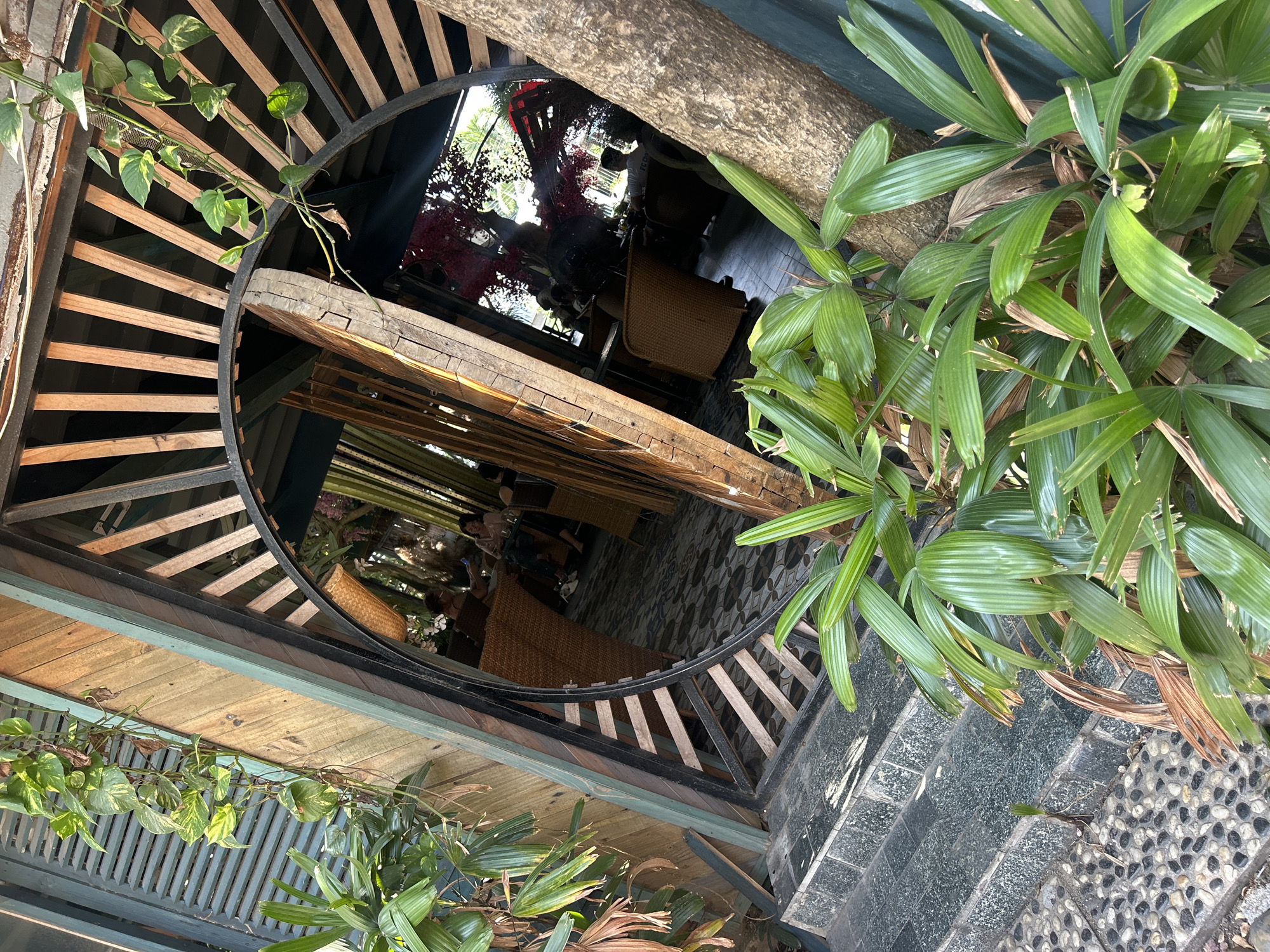 View from above, centered on a large circular opening in a building structure. The opening reveals an interior space with tiled floors, wooden furniture, and people partially visible inside. Lush greenery surrounds the circular opening on the exterior. The photo was taken in Bac Nha Trang Ward, Vietnam.