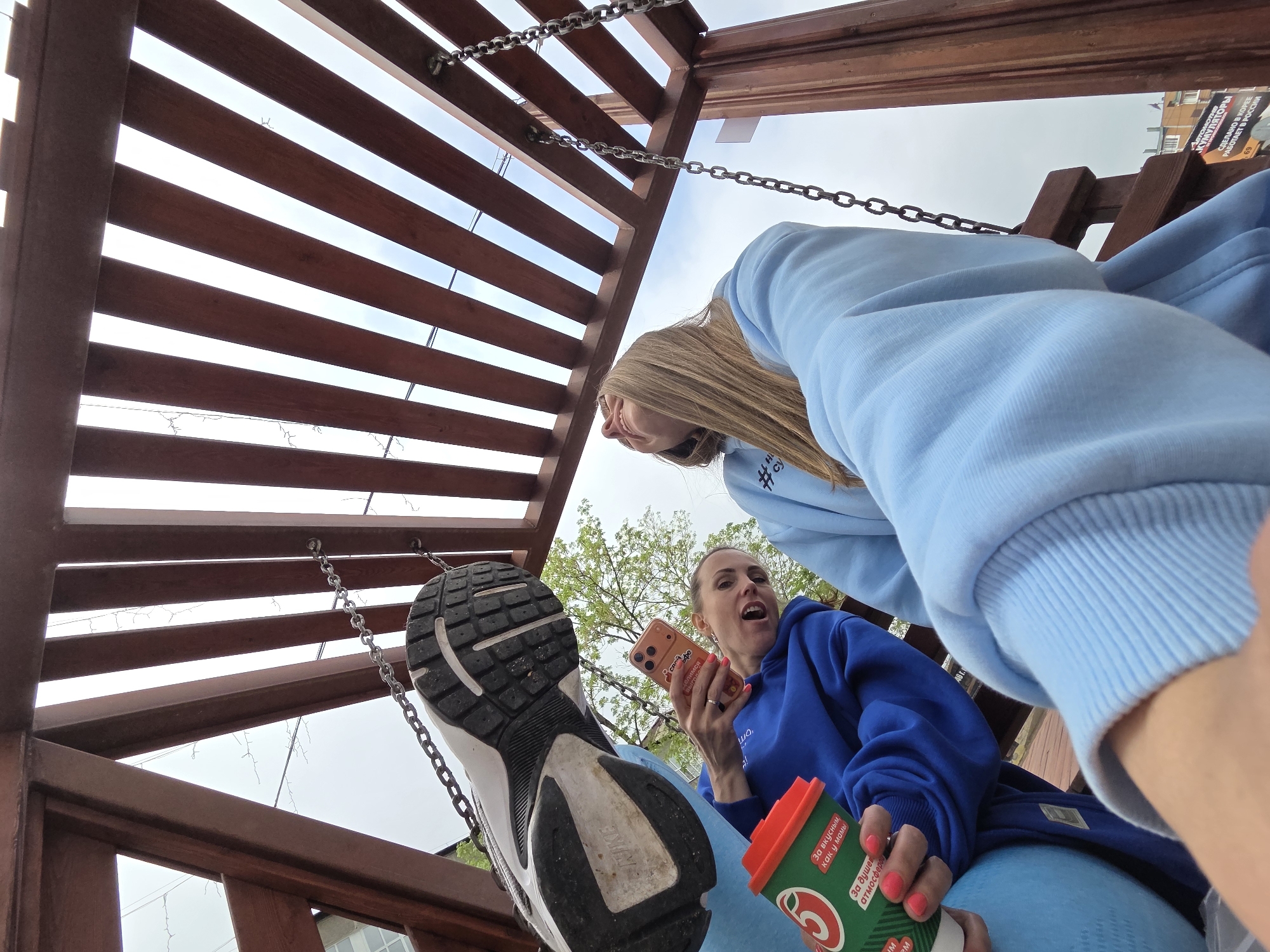 The image, taken from a low angle, captures two individuals on a swing set beneath a wooden structure with horizontal slats. A woman in a blue hooded sweatshirt holds a green and red cup in one hand and a cell phone in the other, her mouth open in an exclamation. Another person, wearing a light blue sweatshirt, is partially visible above and behind her. A sneaker, also part of the scene, hangs from the swing's chain.
The background reveals a cityscape, including buildings and greenery. Based on the Cyrillic text on the cup and a poster visible in the background, the location is identified as Birobidzhan, Russia.