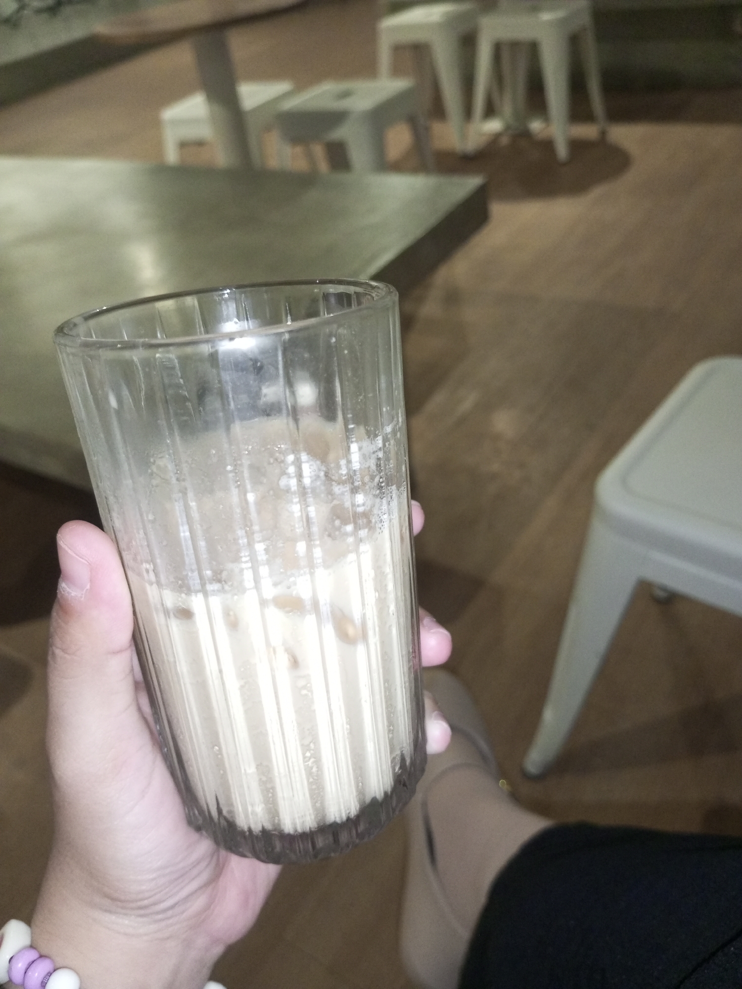 A person is holding a tall glass of a light-brown liquid, possibly iced coffee or a similar beverage, indoors. The glass is about half full with ice visible. The person is wearing a bracelet with purple and white beads on their left wrist and a beige flat shoe can be seen near the bottom. They are wearing black trousers. A table is visible in the background, along with white stools and another white stool is on the right. The floor is wooden. The location is likely a cafe or restaurant. Without additional information or cues, I am unable to determine if the image was taken in North Jakarta, Indonesia.