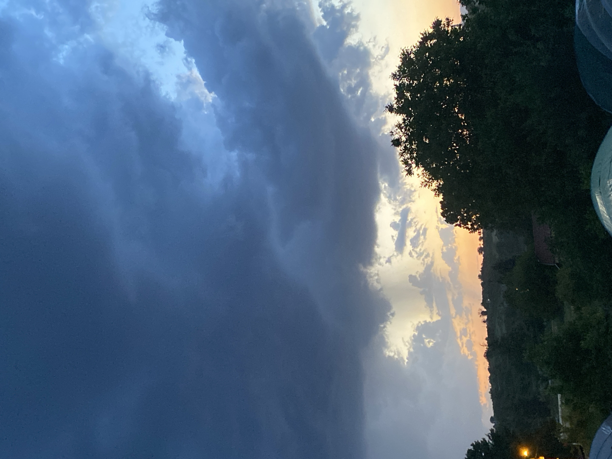 The image depicts a partially cloudy sky at dusk or dawn. Dark, heavy clouds dominate the left side, contrasting with lighter clouds and a golden-orange hue on the right. Trees are silhouetted against the sky on the right edge, indicating a natural environment. The lower right corner shows objects with a blueish-green hue, possibly parts of structures or objects in a yard. There is no clear evidence in the image of the location being Konstantinovo, Bulgaria.