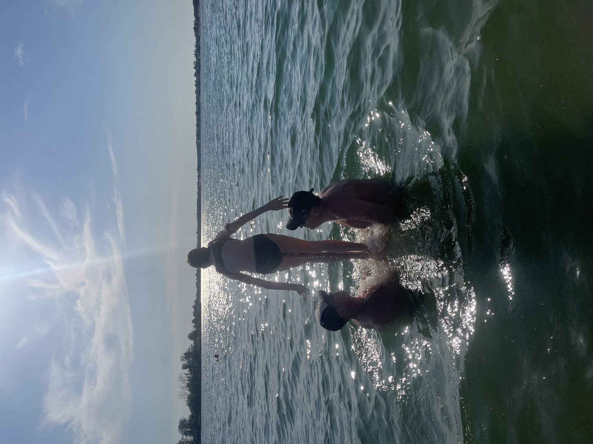 Three people are in the water, partially submerged. The person in the center is wearing a black and white swimsuit and appears to be standing. They are reaching out a hand. To their right, another person is partially submerged, wearing a black baseball cap. To the left of the center person, another individual is mostly submerged, with only their head and shoulders visible. They are also wearing a black baseball cap. The water is choppy and reflects the sunlight, creating many sparkling highlights. In the background, a treeline is visible on the horizon, suggesting a natural body of water. The sky is blue with some wispy white clouds, and the sun is bright, indicating it is daytime, likely late afternoon due to the angle of the sun and the long reflections on the water. The overall scene suggests a recreational activity in a lake or similar body of water. There is no visible text in the image.