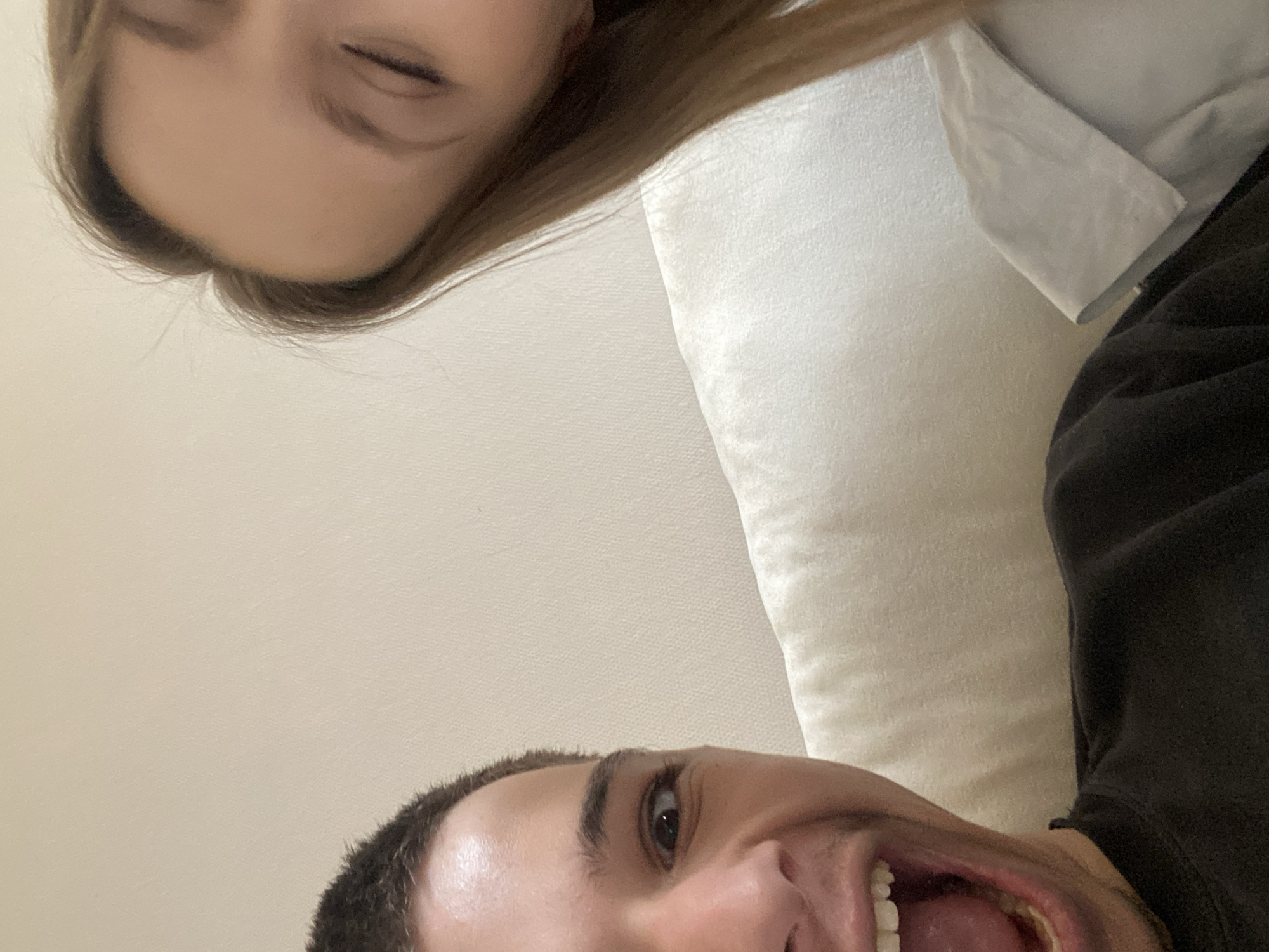 This close-up, low-angle image captures two individuals in a candid, indoor setting. The perspective is from below, looking upwards at their faces, which are oriented diagonally across the frame.
In the upper section of the image, the forehead, closed eyes, and light brown hair of one person, likely female, are visible. Her expression appears calm, suggesting she might be resting or asleep. A white pillow or cushion is partially seen behind her head and a light-colored garment near her left side.
The lower portion of the frame is dominated by the face of another person, likely male, with short dark hair. He has a wide-open eye looking directly towards the camera and his mouth is wide open, revealing his teeth and tongue, suggesting an expression of laughter, surprise, or excitement. Part of a dark garment, possibly a sweatshirt, is visible on his right side, and he also appears to be resting against a white pillow or cushion.
The background is a plain, light-colored surface, likely a wall or ceiling. The lighting is soft and even, indicating an indoor environment. The image conveys a playful or relaxed moment between the two subjects. No specific time of day is discernible from the lighting, and there is no visible text. The implied location, based on the provided context, is Strasbourg, France, though no visual cues within the image confirm this.