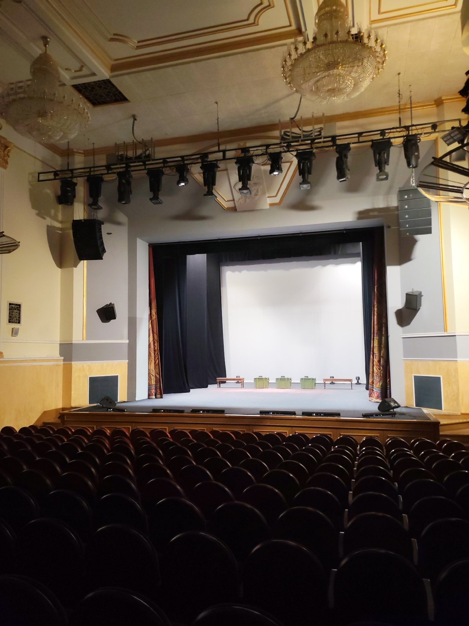 The image displays the interior of an ornate theater or auditorium, viewed from the audience seats looking towards the stage. Rows of dark, empty upholstered seats fill the foreground, creating a sense of quiet anticipation.
The stage is elevated and covered with a light grey or blue carpet. Black curtains are drawn partially to the sides, revealing a large, plain white screen or backdrop at the rear of the stage. Arranged on the stage are several rectangular, light green boxes or seating units, along with a few small wooden benches, suggesting a setup for a presentation, performance, or conference. Small audio monitors are visible on the stage floor at the front.
Above the stage, a metal truss supports numerous theatrical spotlights and lighting fixtures. Large, multi-tiered crystal chandeliers hang from the high, decorative ceiling on both sides of the stage opening. Tall, dark speakers are mounted high on the side walls, while smaller speakers are positioned lower on the paneled walls, which feature cream, grey, and gold accents. A decorative ceiling vent is also visible.
No people are present in the scene. The artificial lighting from the chandeliers and stage lights illuminates the space, suggesting an indoor setting. The grandeur of the venue, with its classical architectural details, points to a historical or cultural performance space, located in Izhevsk, Russia. No legible text is visible in the image.