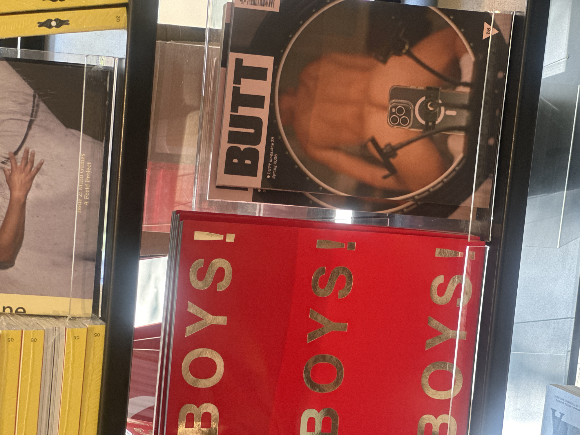 This eye-level shot displays a collection of magazines and books on a shelf, likely in a bookstore in London, United Kingdom. The foreground is dominated by a red book with the word "BOYS!" printed in gold lettering on its cover multiple times. Adjacent to this, there's a magazine titled "BUTT" featuring a photograph of a torso being filmed with a mobile phone. To the left, there are publications with yellow spines and covers showing a hand. The shelf itself is black and constructed of clear acrylic, which allows light to filter through, with elements of the environment visible in the blurred background. The lighting is bright and even, highlighting the colors and textures of the items on the shelf.