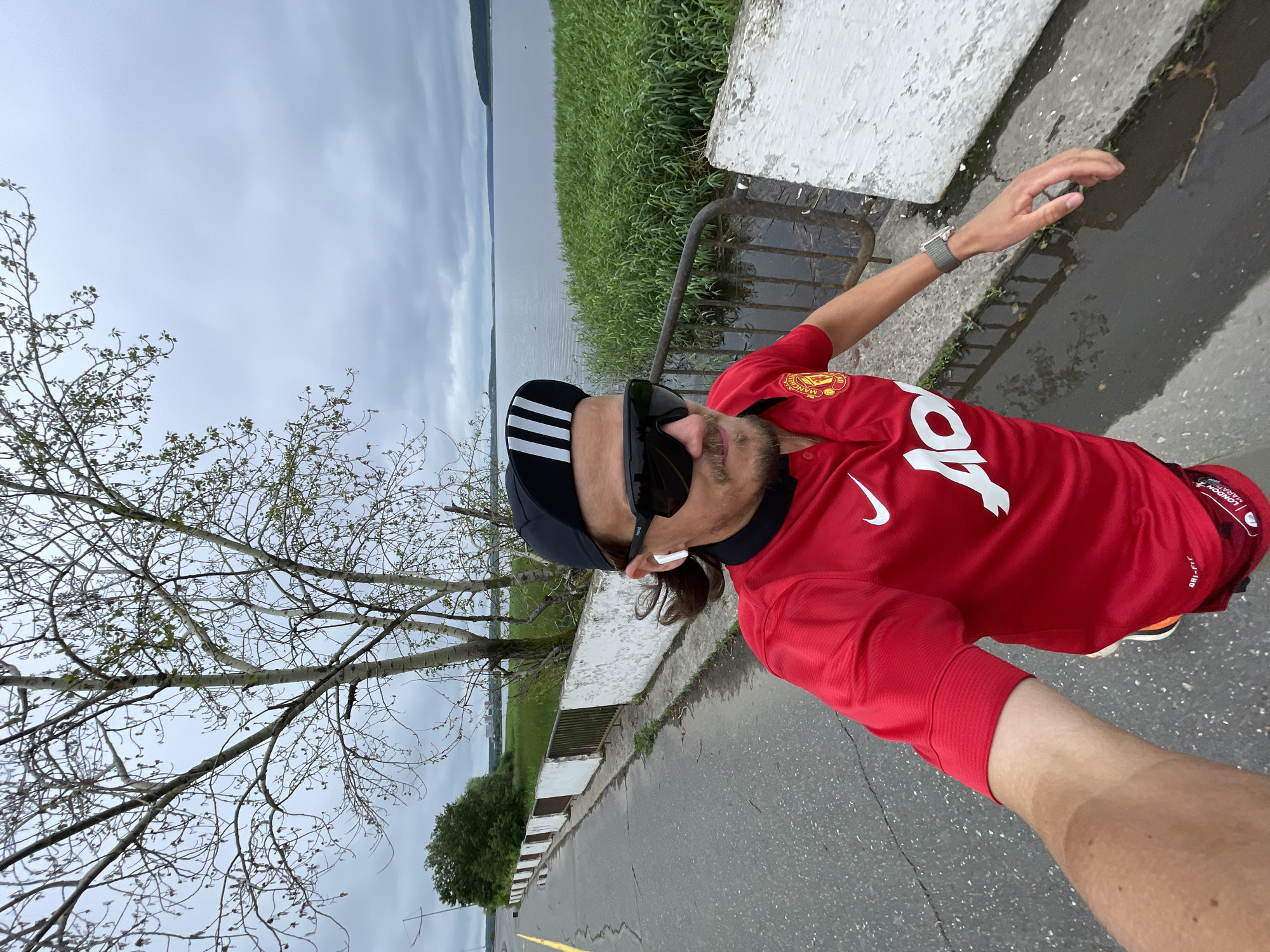 A man wearing a red Manchester United jersey, black baseball cap, and sunglasses is running along a road next to a body of water. The sky is overcast, and the road is wet, suggesting it may have recently rained. Green grass and reeds grow along the edge of the water. There is a low white wall and a metal railing separating the road from the water. Trees with sparse leaves are visible in the background.