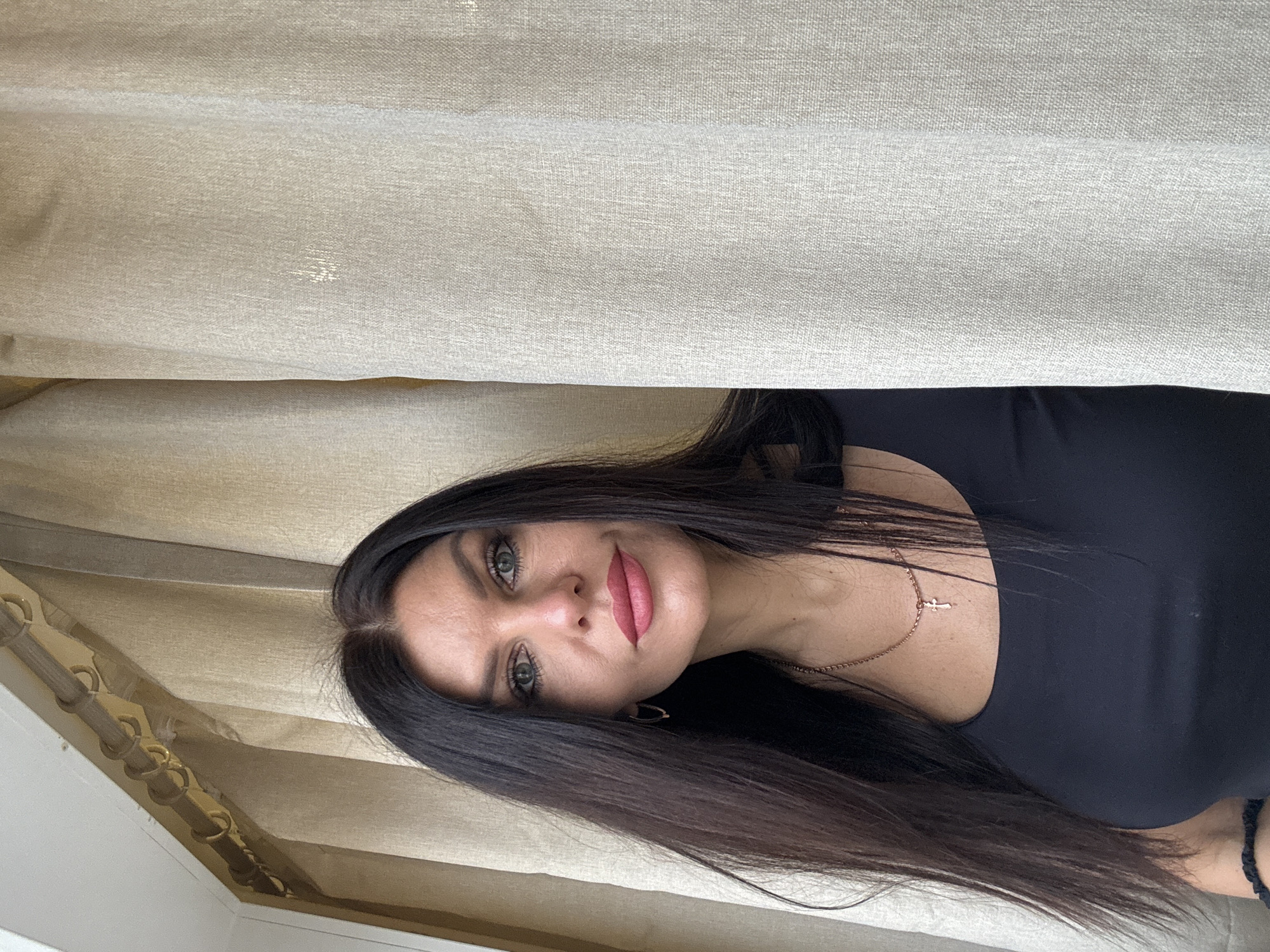 The image is a medium close-up portrait of a woman, framed from the chest up against a backdrop of light-colored curtains. The original image is oriented sideways, but the description assumes a corrected upright view.
The woman has long, straight, dark brown or black hair that frames her face and falls over her shoulders. She has striking light-colored eyes, possibly green or hazel, and her lips are adorned with pinkish-red lipstick. She is looking directly at the camera with a calm and gentle expression, featuring a subtle smile. Her skin tone is fair.
She is wearing a dark-colored top, possibly black, with a rounded neckline. Around her neck, she wears a delicate, rose gold-toned chain necklace with a small cross pendant. A dark, possibly braided, bracelet is partially visible on her right wrist.
The background consists primarily of light beige or off-white curtains, which appear to be made of a textured, woven fabric, showing subtle vertical folds and some areas of highlights. On the upper left side of the frame, a light brown curtain rod with multiple curtain rings is visible, suggesting an indoor setting. The lighting appears soft and natural, possibly coming from behind or through the curtains, creating gentle illumination on the subject and the fabric.
The image captures a posed portrait of the woman. There is no visible text in the image. The implied setting is indoors in Chaikovsky, Russia.