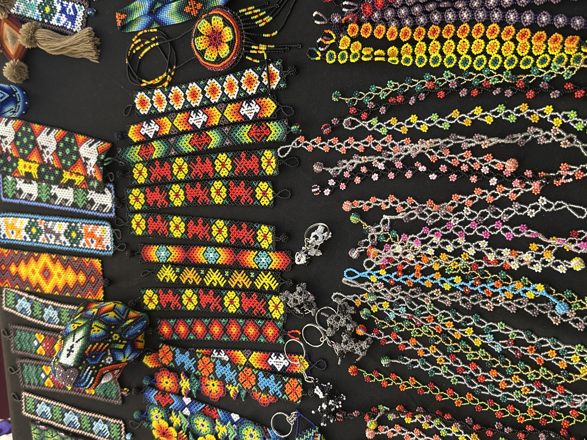 The image presents a diverse collection of beaded crafts meticulously arranged on a black, flat surface, indicative of a marketplace or retail display in Mexico City, Mexico. In the upper portion, several long, rectangular beaded bands, likely bookmarks or decorative elements, are displayed horizontally. These feature intricate patterns, including geometric designs, stylized animal figures, and abstract motifs, rendered in vibrant hues of yellow, orange, red, green, blue, white, and black. Centrally located among these bands is a three-dimensional, multi-colored beaded object, possibly a sculpture or container, exhibiting a complex mosaic of blue, green, white, orange, and red beads. The upper right corner shows a small blue beaded pot, two brown yarn tassels, and a beaded pendant on a black cord.
Below this, a horizontal row of narrower beaded bracelets is visible, predominantly featuring repeating floral patterns in red, yellow, and orange, often set against black, green, or white backgrounds. To the far left, a wider bracelet displays a complex floral design incorporating blue, green, yellow, and red beads. Adjacent to these, on the right, is a circular beaded pendant with a central orange, yellow, and red floral motif, suspended from a black beaded necklace with multiple dangling strands.
The lower half of the image is filled with numerous delicate beaded chains, likely necklaces or bracelets. These are composed of small, individual beads, frequently forming tiny floral clusters or continuous strands in a broad palette of colors including red, orange, yellow, green, blue, pink, white, black, and silver. They are arranged in parallel or overlapping lines, creating a dense, colorful texture. On the far right, wider beaded bands are present, densely packed with circular or flower-like motifs in solid, graduated rows of yellow, orange, red, green, blue, and purple. In the mid-left section, several beaded keychains are scattered, featuring silver metal keyrings attached to small, three-dimensional beaded figures in grey and white, alongside a blue star-shaped charm. No individuals are present in the scene.