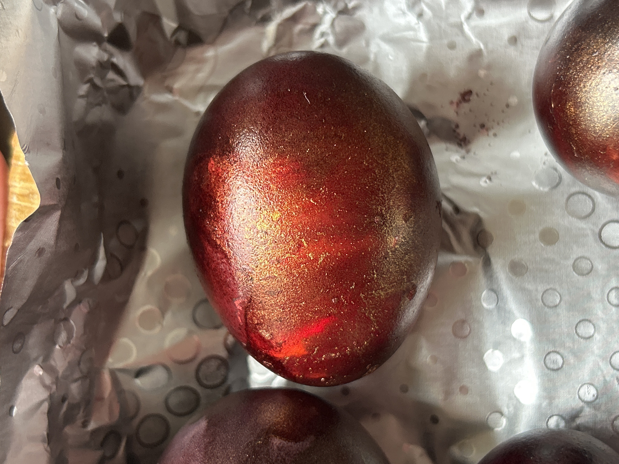 Several metallic, red and gold colored eggs are resting on a textured, silvery surface. The surface appears to be patterned with embossed circles. The background is out of focus. There is no indication of the location being Krumovo, Bulgaria. No people, actions, or interactions are visible.