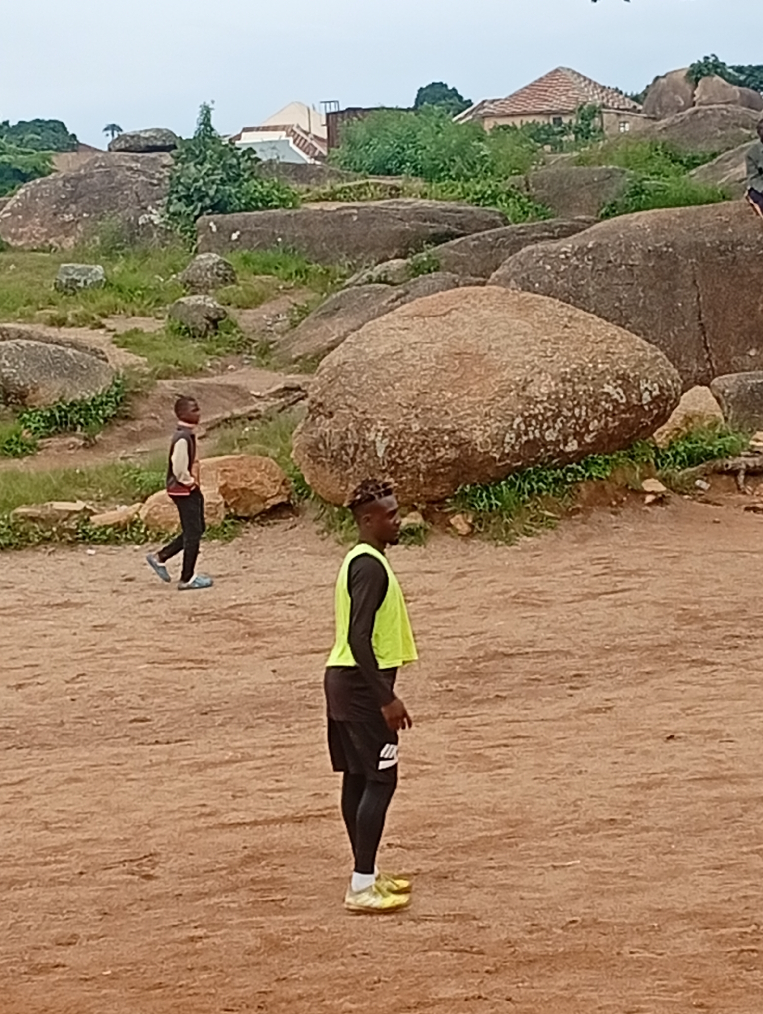 In an outdoor, rocky landscape, two young males are visible. The male in the foreground, facing to the right, is wearing a bright yellow vest over a black t-shirt and black shorts. He has on black leggings and yellow athletic shoes. The second male, positioned further back and to the left, is walking away from the camera. He is wearing a jacket with white and red accents, black pants, and blue shoes. The terrain consists of large boulders, patches of grass, and dirt. In the background, a building with a red tiled roof is situated amongst the rocks and vegetation. The sky is overcast.