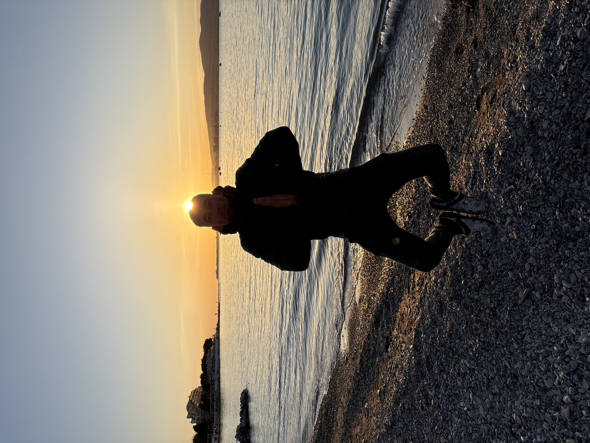 The image captures a serene coastal landscape at sunset, with a person standing on a pebbly beach. The scene is set in Gelendzhik, Russia.
In the foreground, a person is silhouetted against the bright, low-hanging sun, which creates a radiant halo around their head. They appear to be an adult, dressed in dark clothing, possibly a hooded jacket and trousers, with lighter-colored footwear. The person is striking a pose, with hands clasped together in front of their chest, suggesting a moment of contemplation, meditation, or a specific posture like a greeting.
To the left of the person is the calm sea, stretching into the distance. The water reflects the warm golden light of the setting sun, creating shimmering patterns on its surface. Waves gently lap the pebbly shore, visible on the right side of the frame, which is covered with various shades of grey and brown stones.
The sky exhibits a clear gradient, transitioning from a brilliant orange and yellow near the horizon where the sun is positioned, to a soft blue higher up. In the far distance, across the water, landmasses, possibly mountains, are visible as faint silhouettes. Further along the coastline to the left, outlines of distant buildings or a townscape can be seen. The overall lighting indicates late afternoon or early evening, characterized by warm, soft light and long shadows, contributing to a tranquil and picturesque atmosphere. There is no visible text in the image.