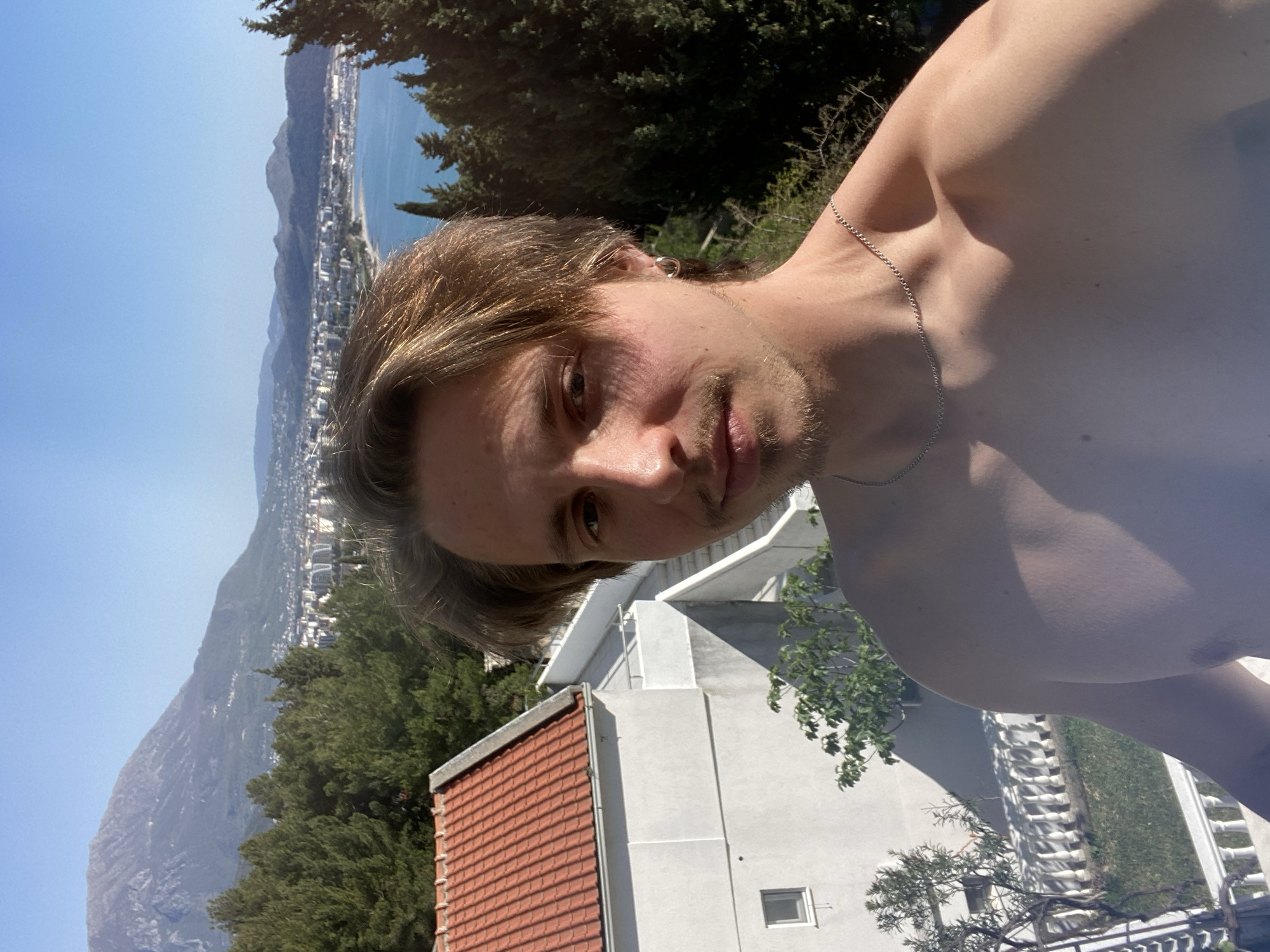 The image shows a shirtless man with light brown hair and a stubbled beard, wearing a silver chain, against a background of Šušanj, Montenegro. The man is positioned close to the camera, filling much of the frame. Behind him is a hillside with dense green trees, a building with a red tile roof and a white exterior. Further in the distance is the coastline with a town and mountains, receding into the horizon under a clear blue sky. The image is rotated, with the landscape appearing to be on its side relative to the man.