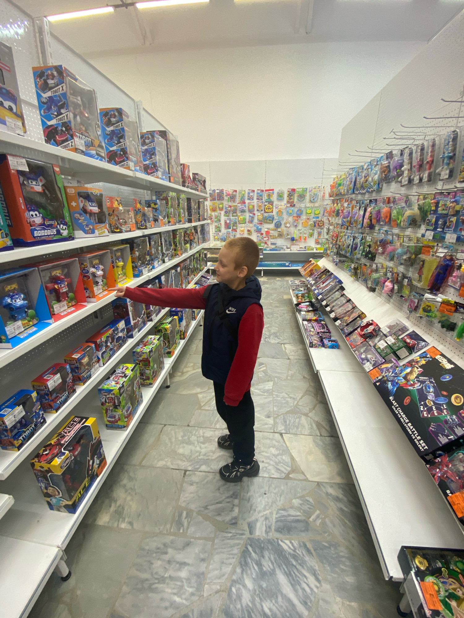 The image shows a young boy in a toy store. The boy is wearing a red and black jacket and black pants, and he is reaching towards a shelf of toys. The shelves are stocked with various toy boxes, many of which appear to be action figures or robots. The store has fluorescent lighting and tiled flooring. The context provided specifies the location as Chaikovsky, Russia, indicating this is a retail environment within that city. The boy is the sole subject visible in the foreground.