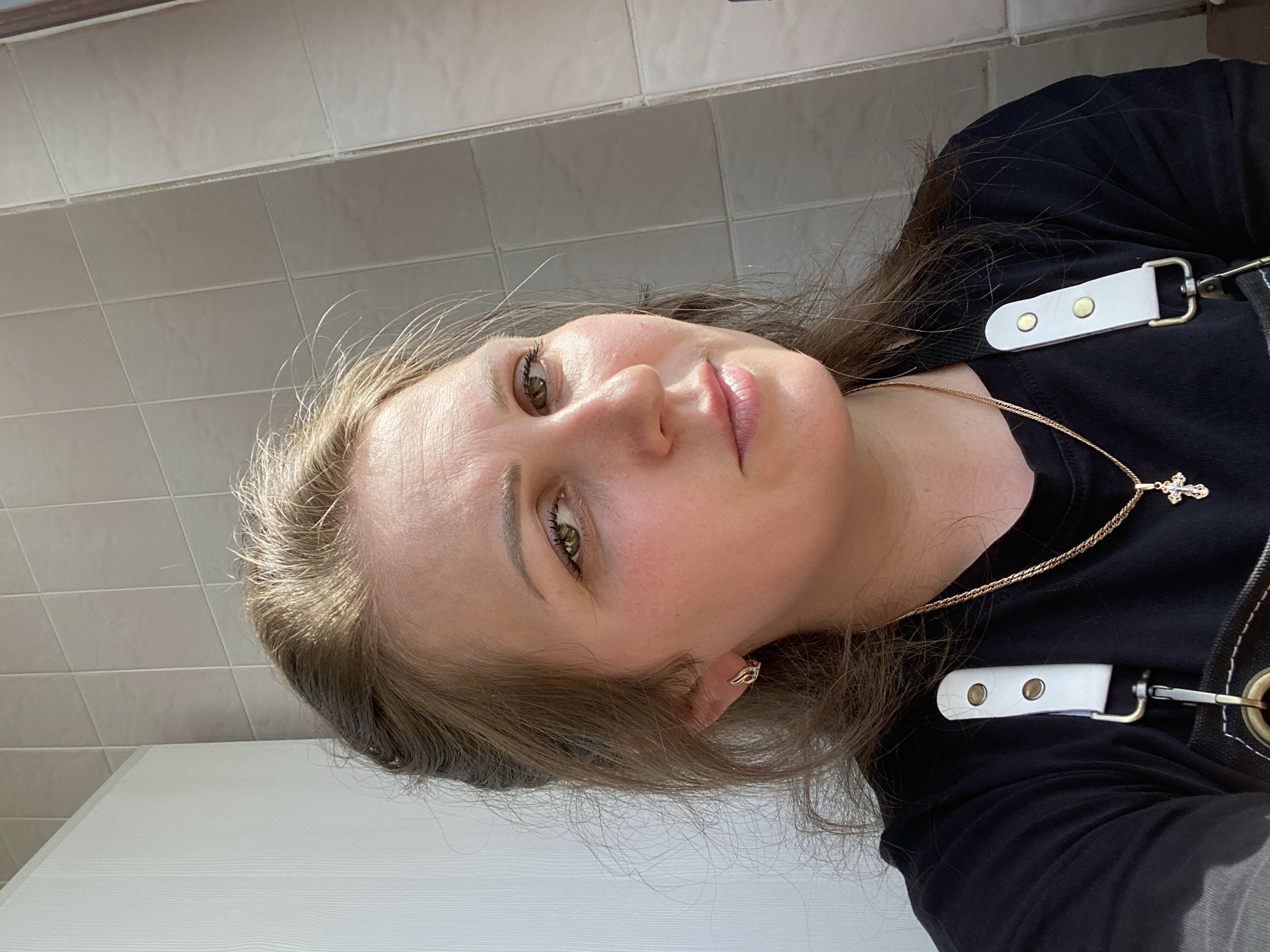 A woman with fair skin and light brown hair is positioned in front of a tiled wall and a white, paneled surface. She has light eyes and pink lips. She is wearing a black shirt with white straps secured by metal buckles, and a gold necklace with a cross pendant. A small gold earring is visible in her ear. The location appears to be indoors. There is no specific evidence to suggest the exact location in the city of Марковский, Russia.