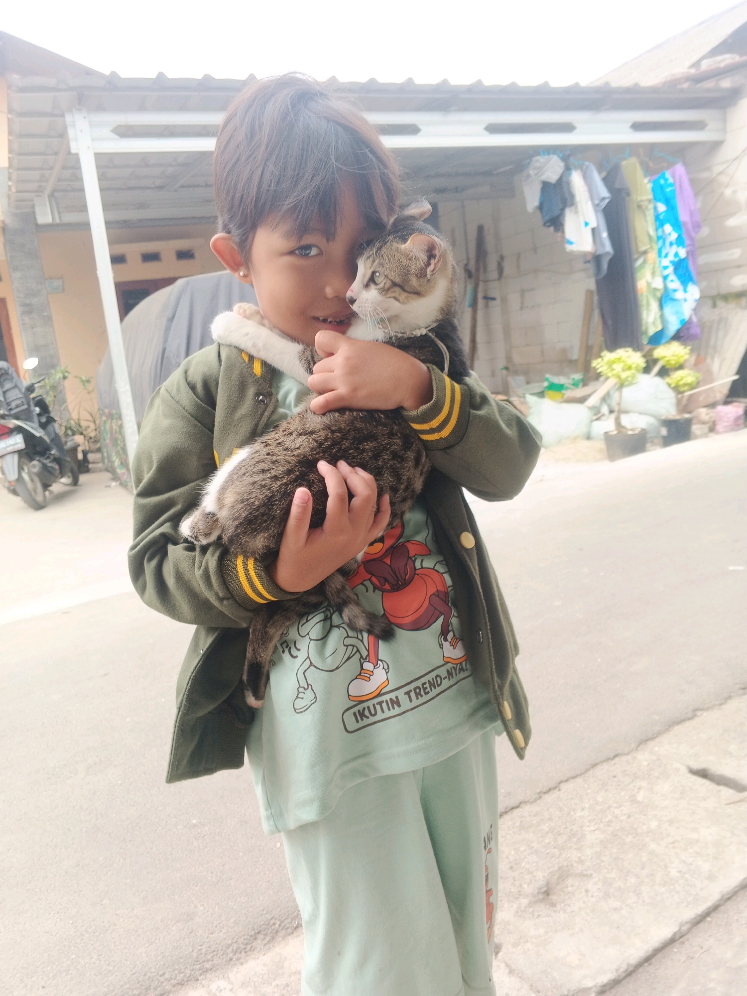 In a residential outdoor setting in Kadu, Indonesia, a young child with dark hair, wearing a green jacket over a light blue t-shirt with a cartoon graphic and text, embraces a tabby cat. The child's face is turned towards the viewer, and they are smiling. The cat is held securely in the child's arms, with its head peeking over the child's shoulder. In the background, a roofed structure with clothes hanging on a line is visible, along with some potted plants and a motorcycle parked to the left. The ground appears to be paved with asphalt and concrete.